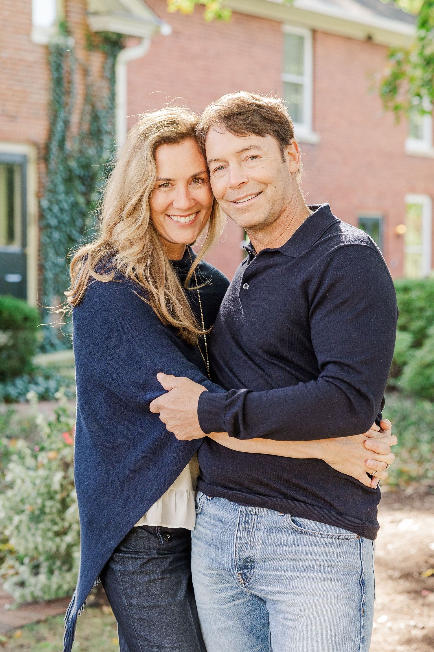 Happy couple in matching navy sweaters embracing outside brick building with garden view.