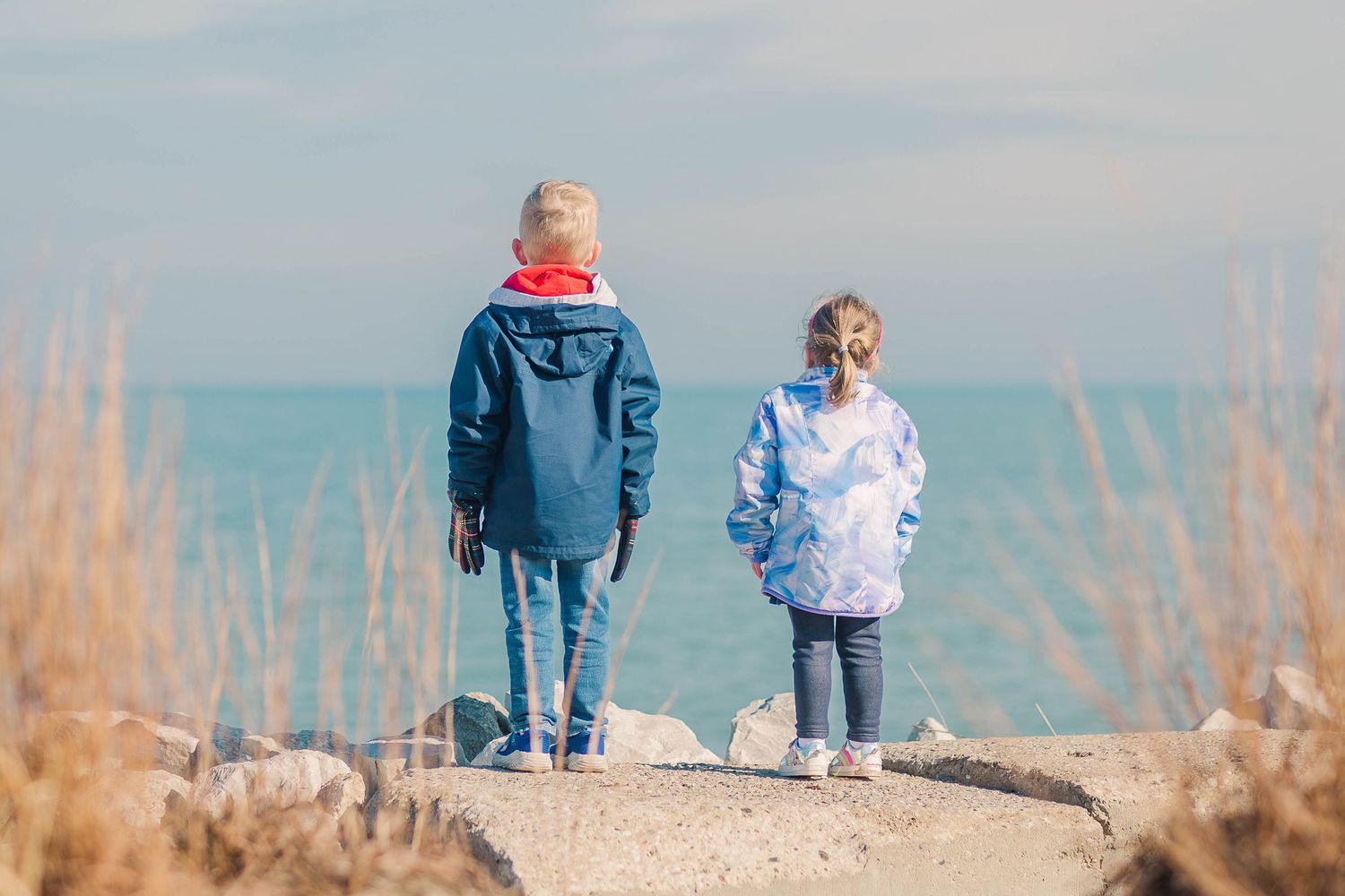 Two figures in winter coats standing on coastal path overlooking the ocean.