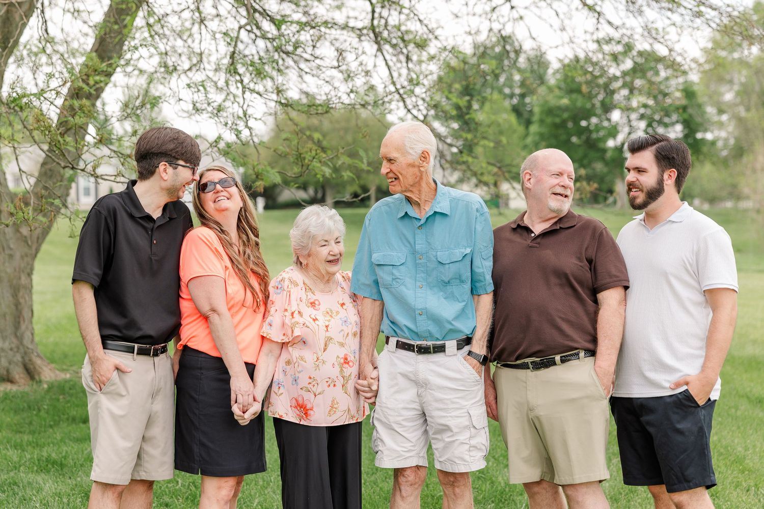 Multi-generational family of six smiling together outdoors on green grass under a tree during a family portrait session.