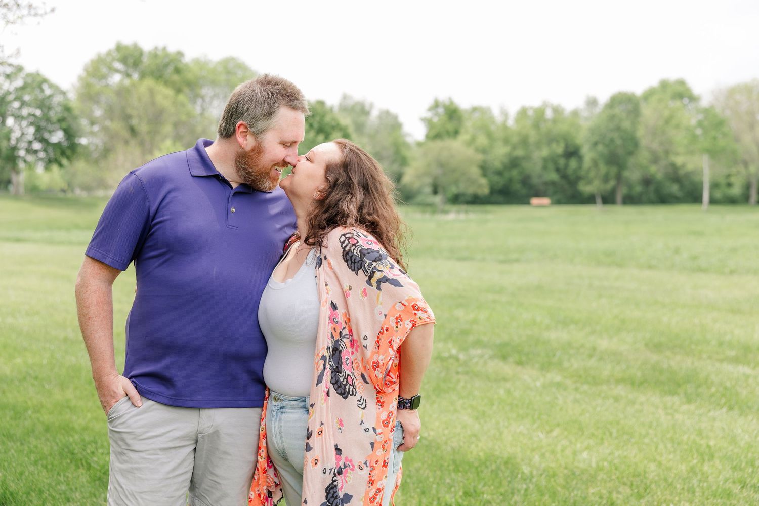 Couple shares a kiss while standing in an open green field during an outdoor family photography session.