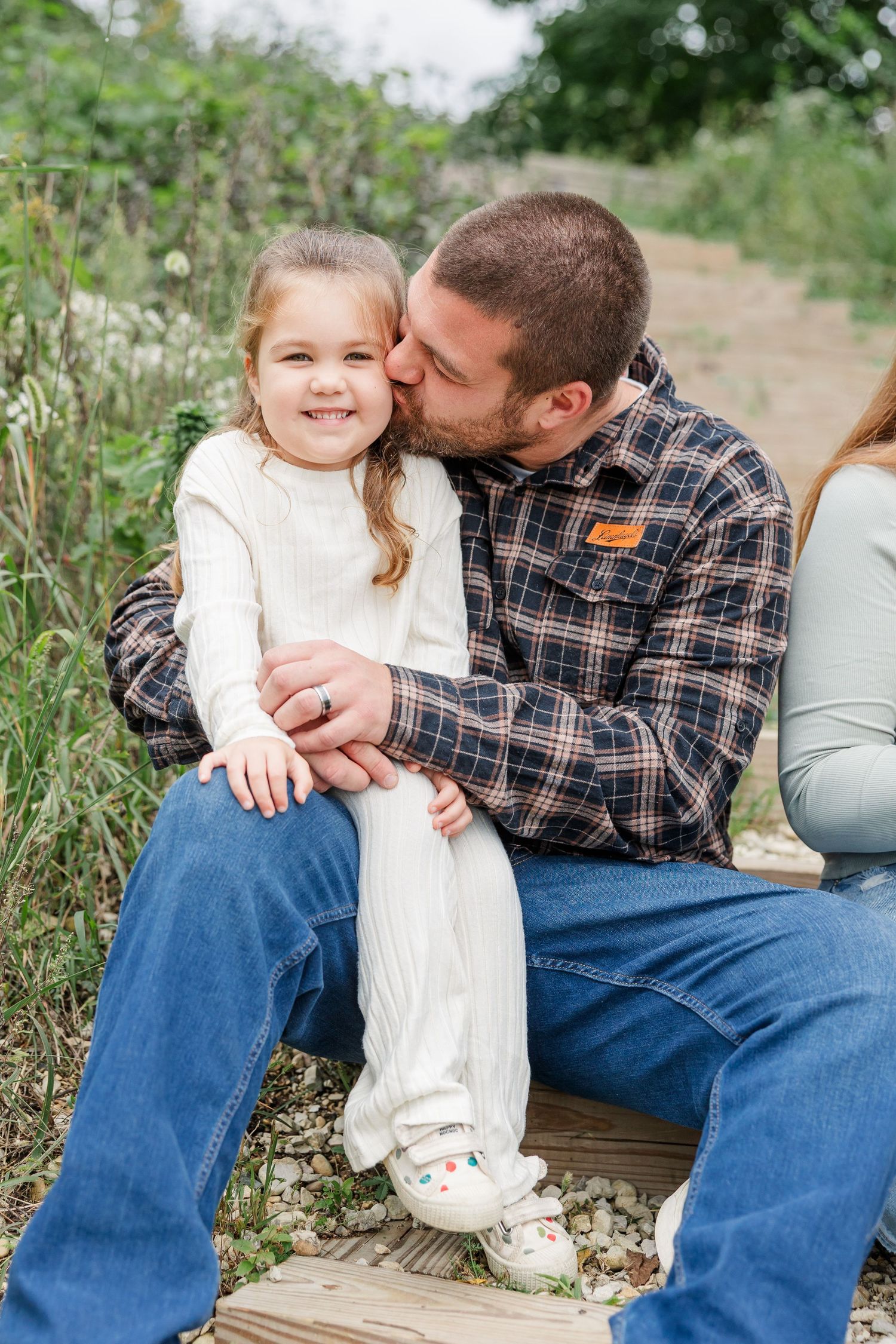 A father kisses his smiling young daughter on the cheek while sitting outdoors on a wooden path.