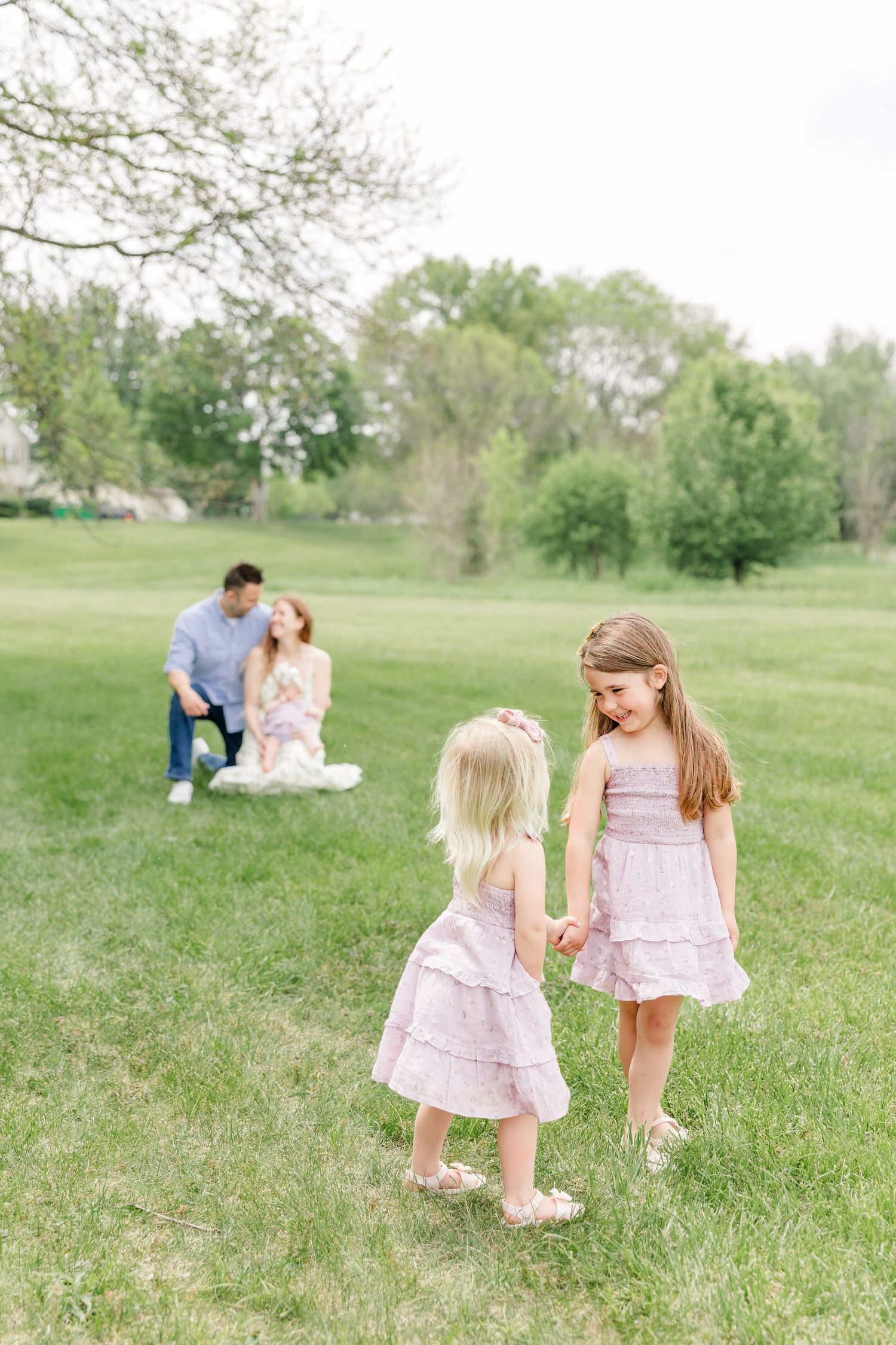Two young girls in pink dresses hold hands in a green park while their parents sit together in the background.