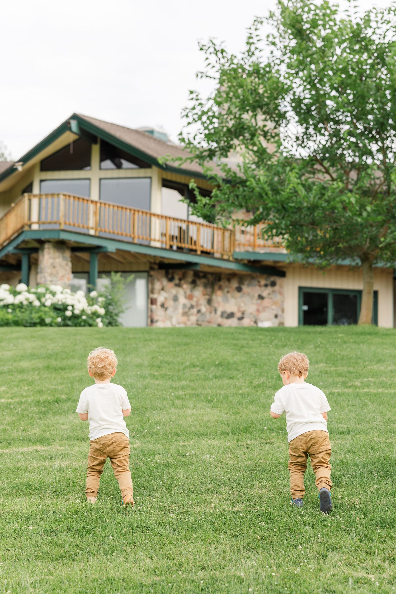 Two young toddler boys in matching white shirts and tan pants run across a green lawn toward a large house.