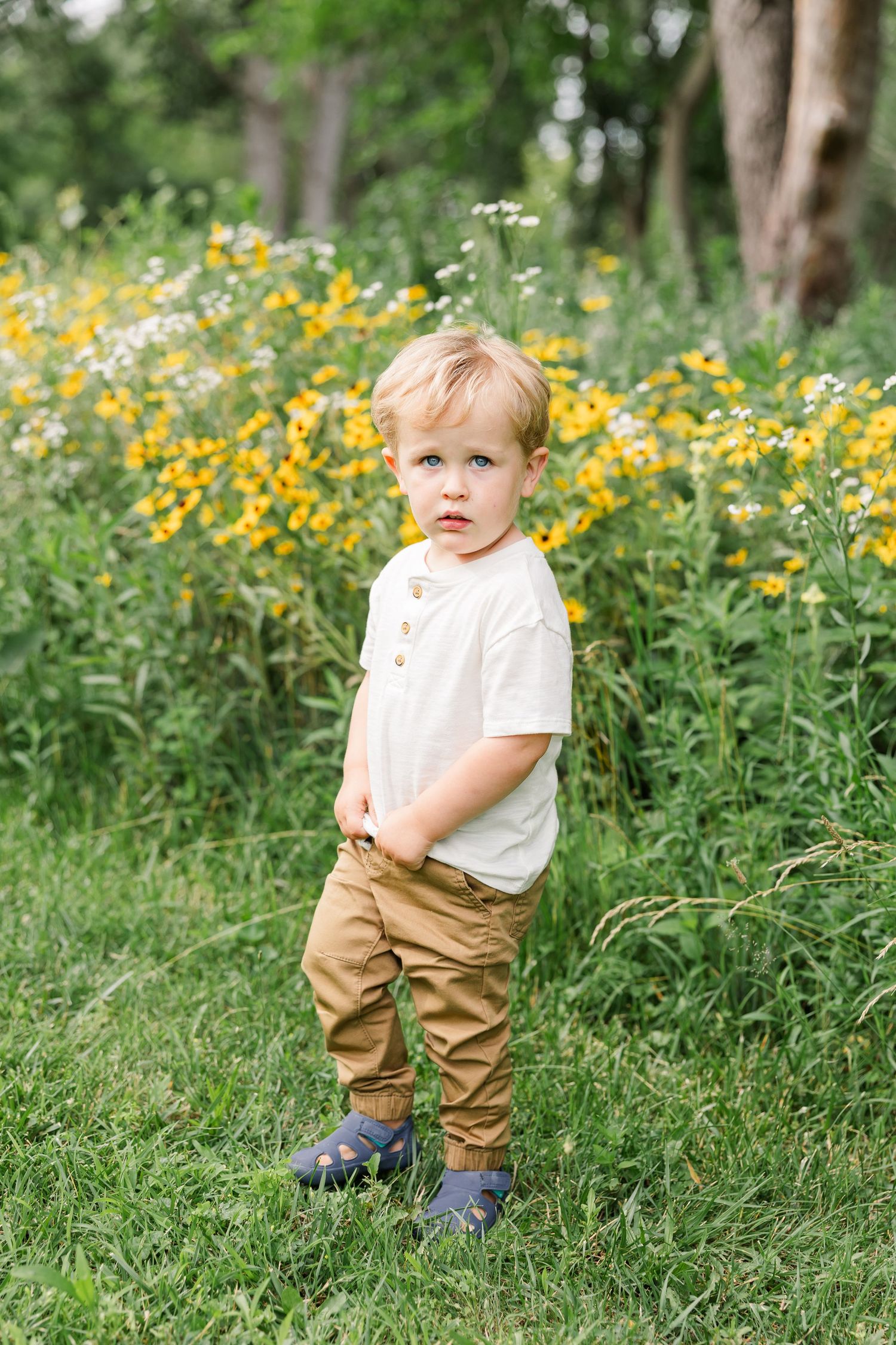 Toddler boy in white shirt and tan pants standing in a green field with yellow wildflowers blooming behind him.
