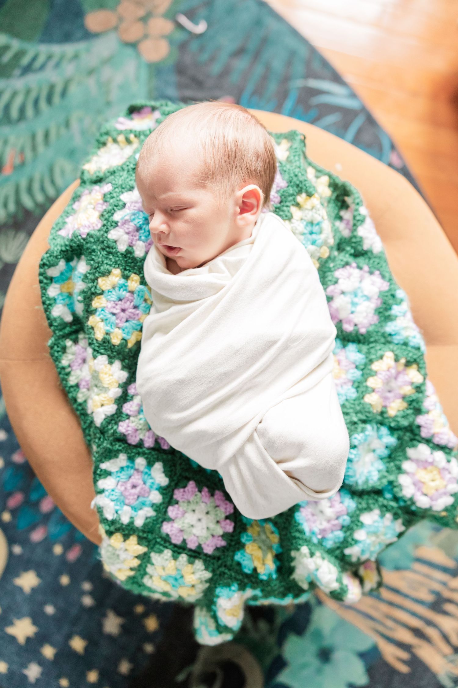 Swaddled newborn baby sleeping peacefully on a colorful crocheted granny square blanket viewed from above.