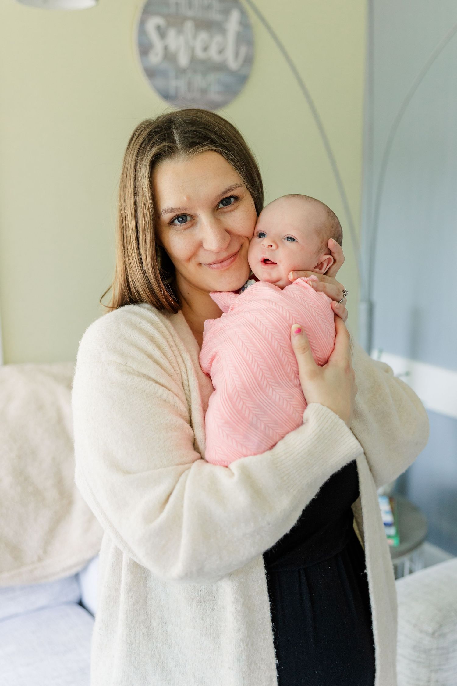 A young mother in a white cardigan tenderly holds her newborn baby wrapped in pink in a cozy home setting.