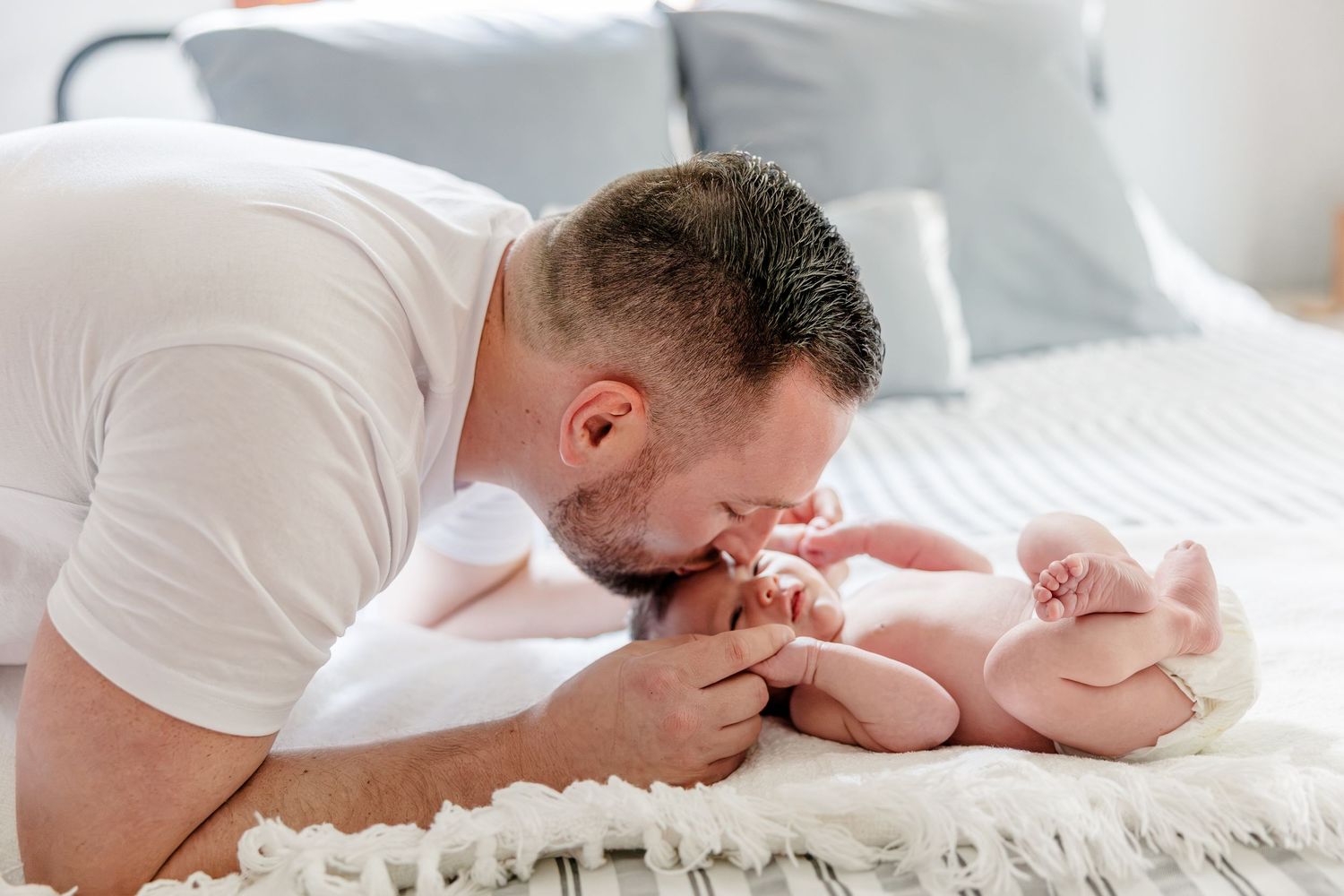 A loving father gently kisses his newborn baby lying on a white blanket on a bed.