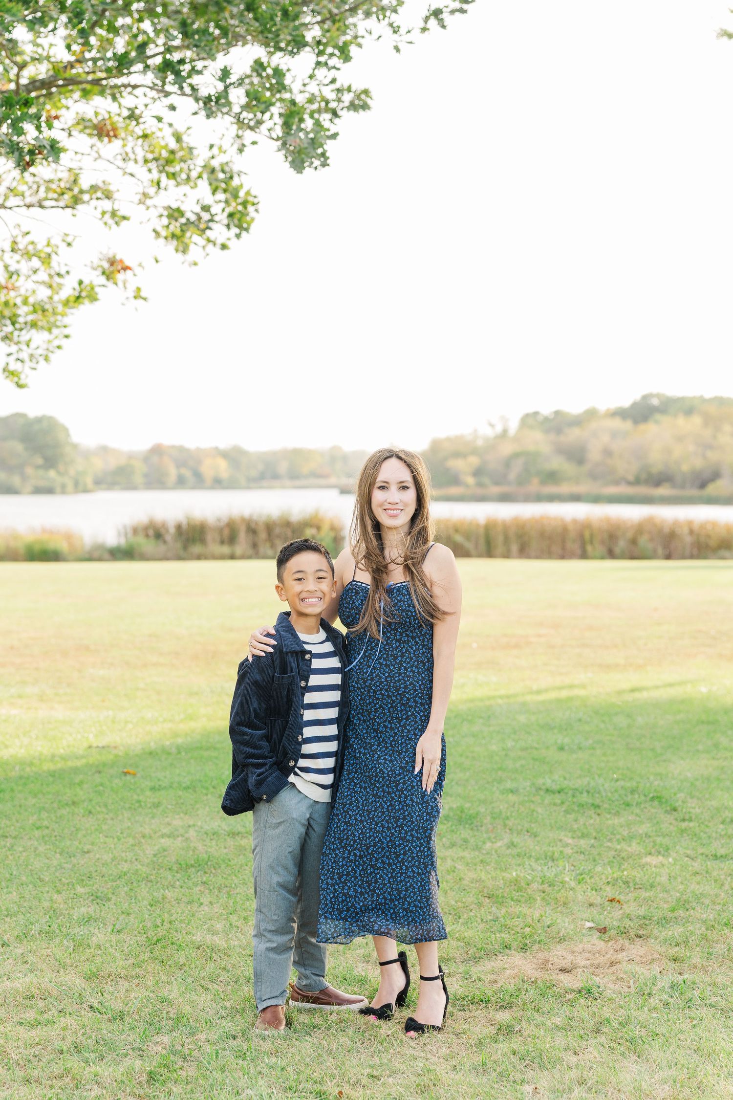 Two people standing together on grassy field with lake and autumn trees in background on sunny day. Busse Woods