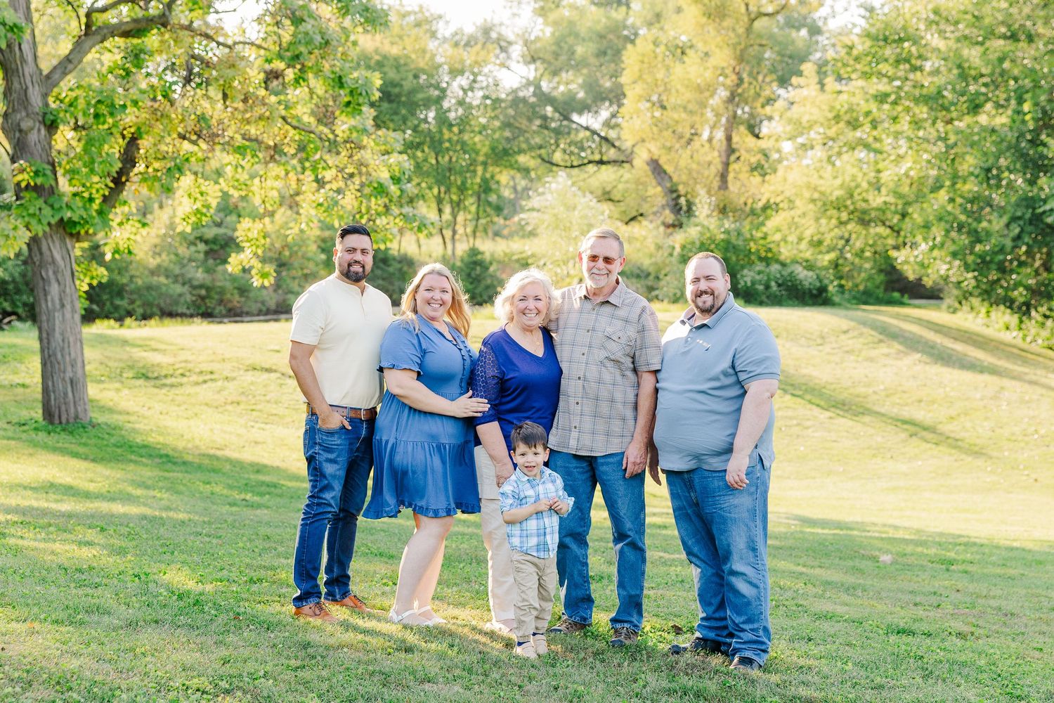 A group of people in coordinated blue and gray outfits posing together in a sunny grassy field during golden hour.Sorg family portraits at S