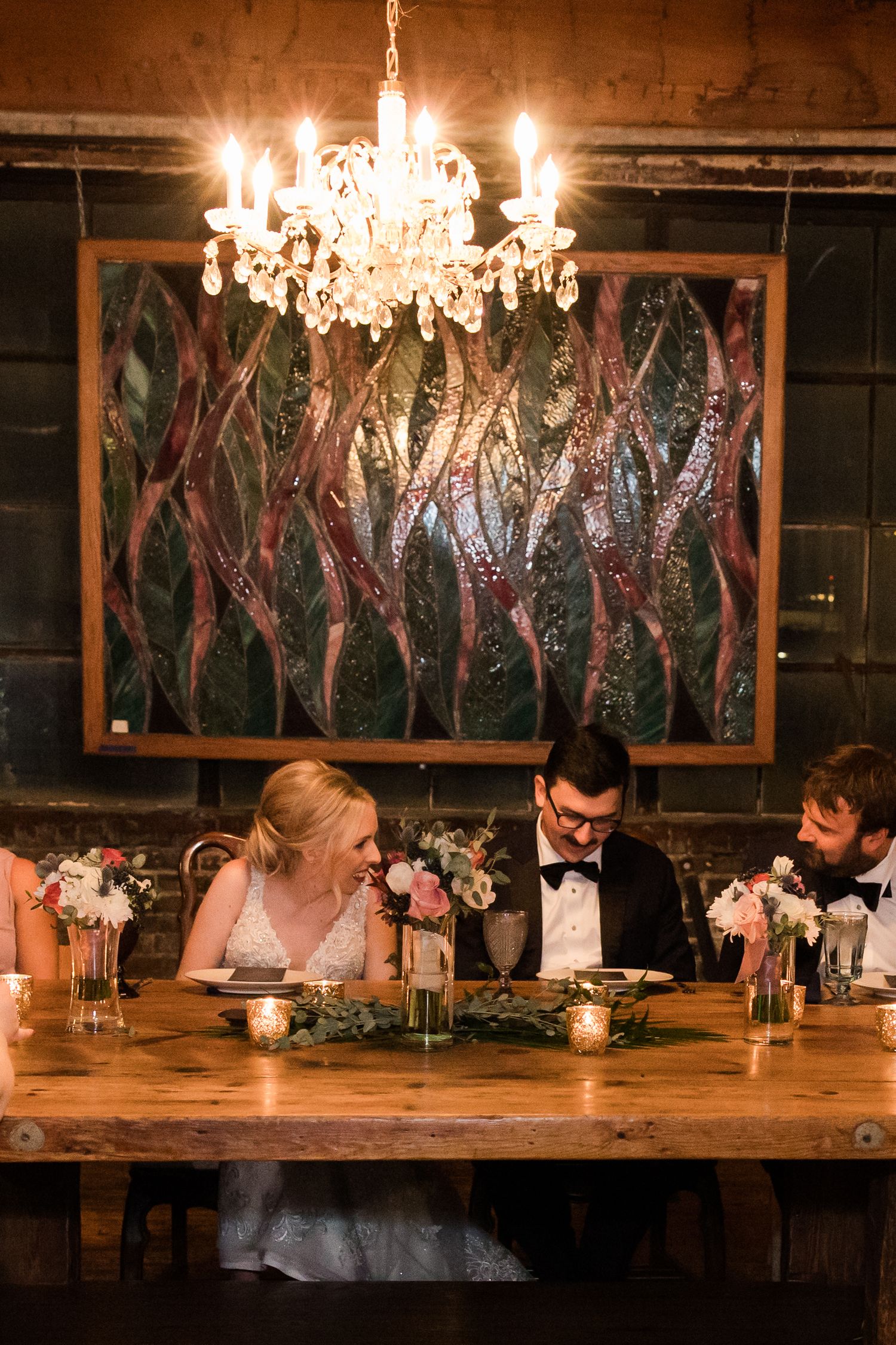 Elegant chandelier illuminates wedding reception table with artistic backdrop and floral arrangements.