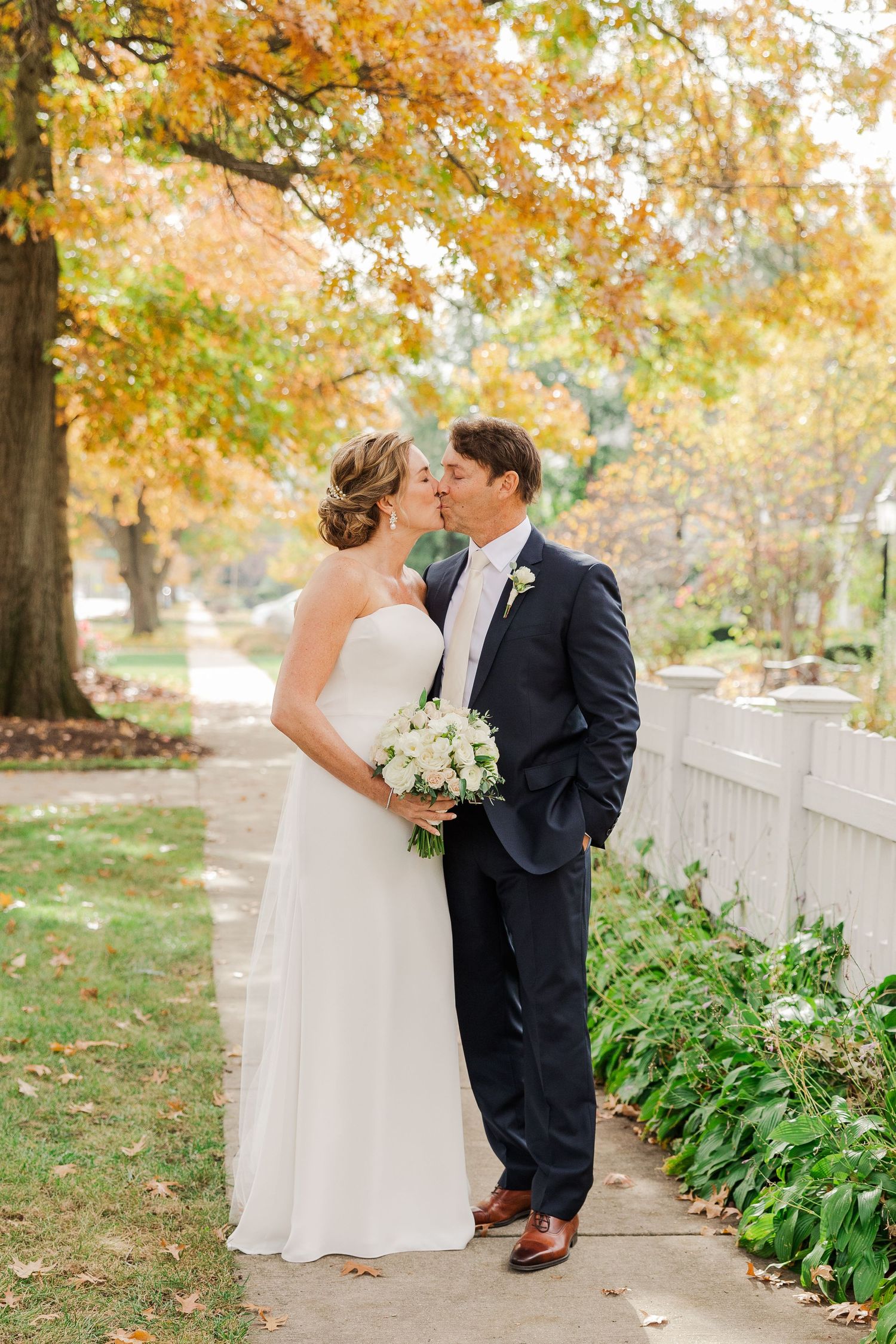 A romantic autumn wedding photo on a garden path with white picket fence and golden leaves.