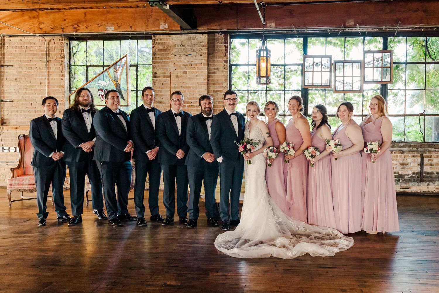 Wedding party lines up in formal attire with bridesmaids in pink dresses and groomsmen in black tuxedos.
