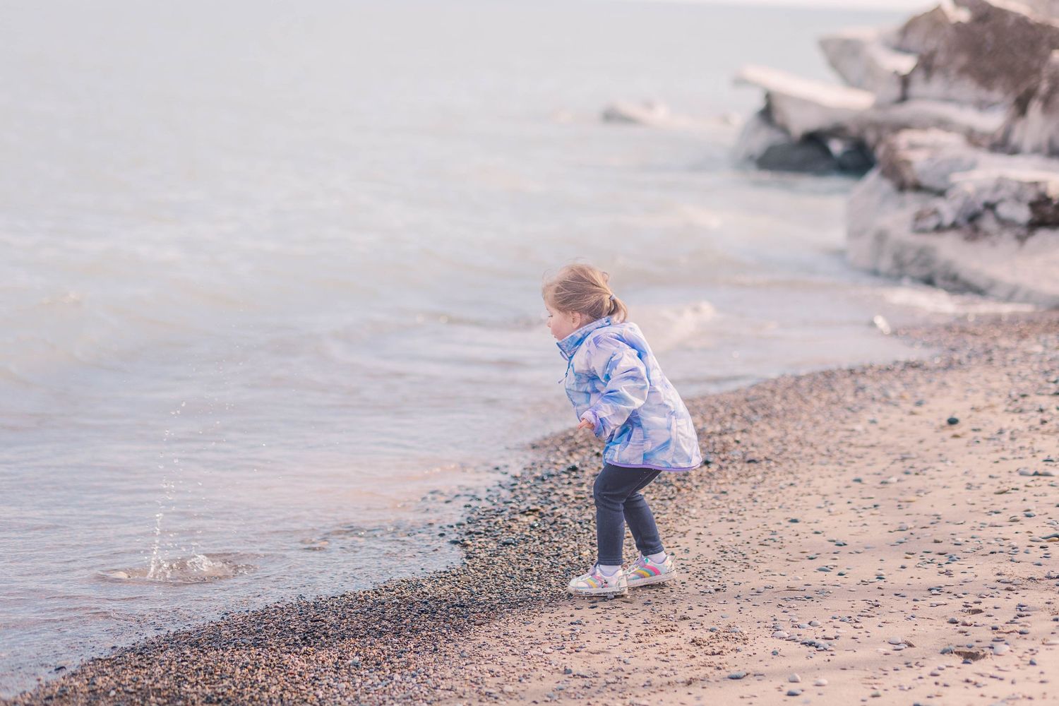 Child in light colored jacket explores rocky shoreline on cloudy day.