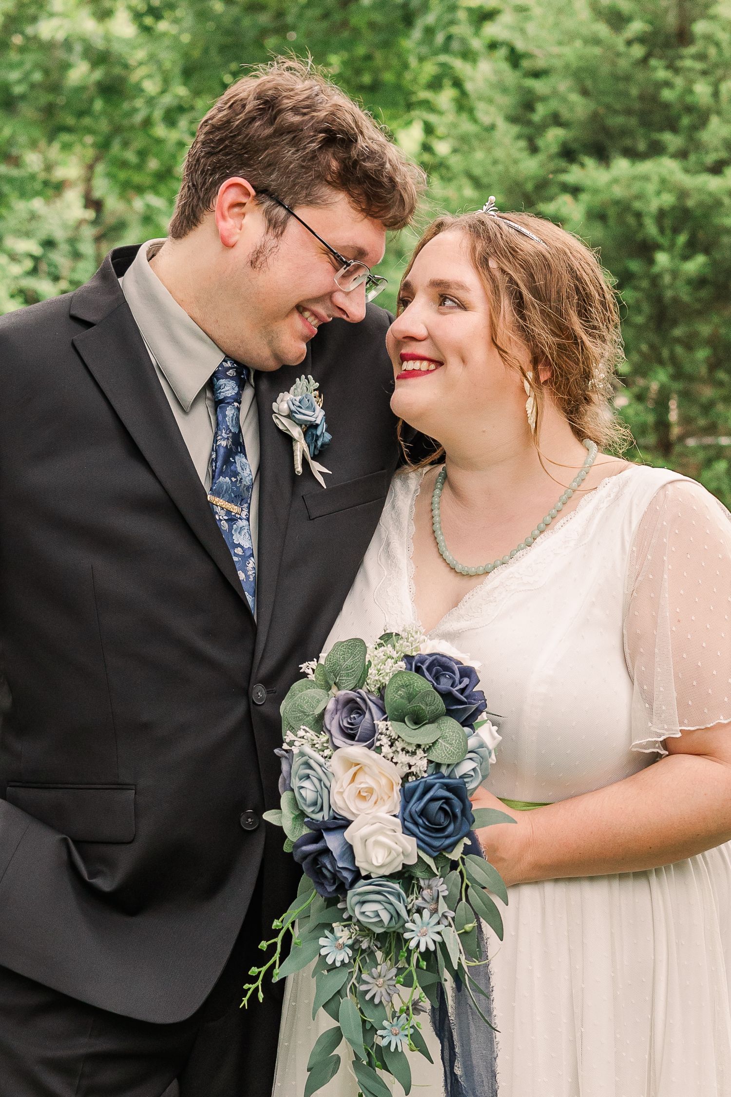 Joyful wedding portrait with blue and white bouquet against green foliage.