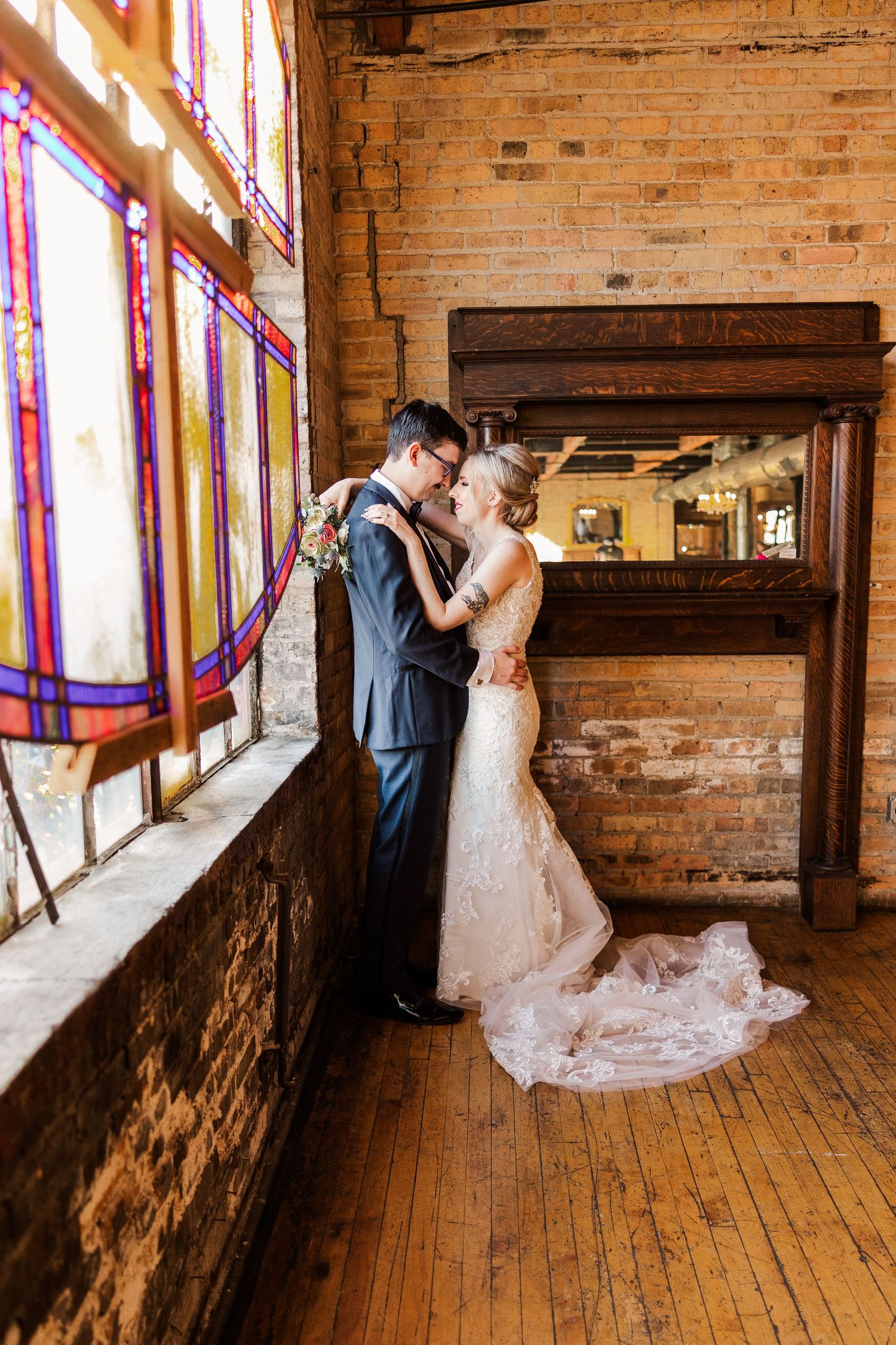 A wedding couple embraces by colorful stained glass windows in an industrial-style venue.