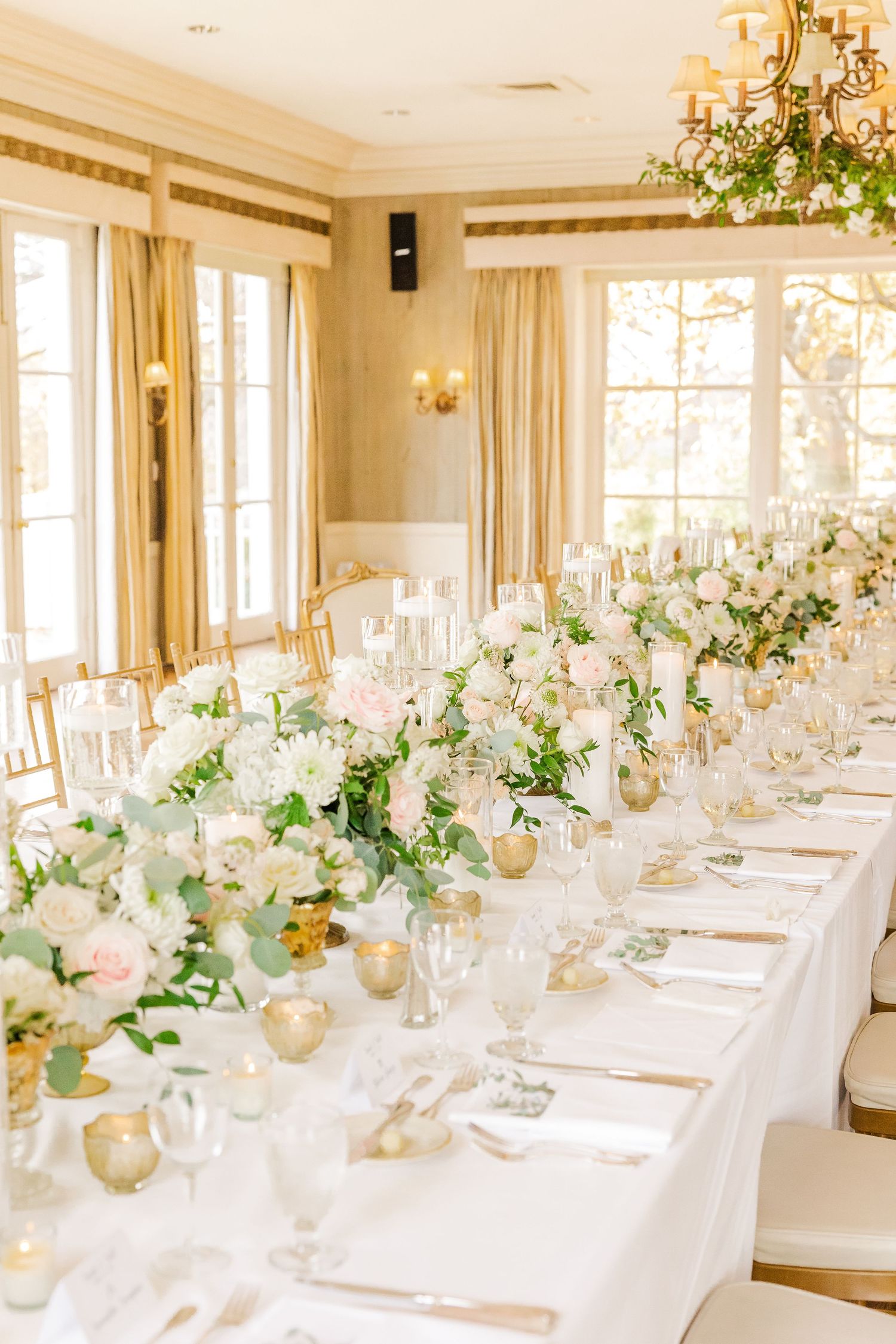 Sun-filled reception room with long table decorated in white and blush floral centerpieces and gold accents.