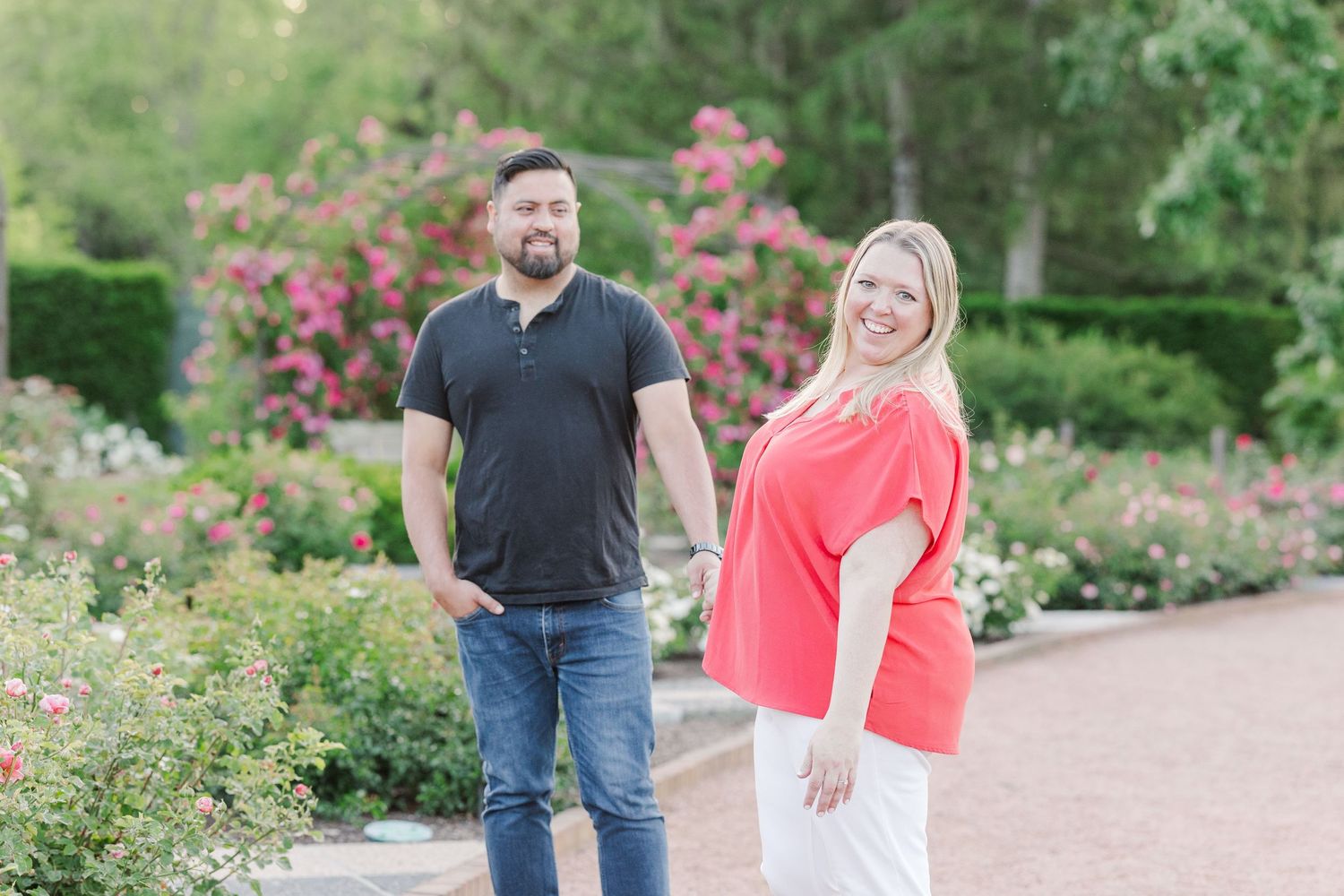 Couple standing casually in a lush garden setting with flowering bushes and pink blooms.