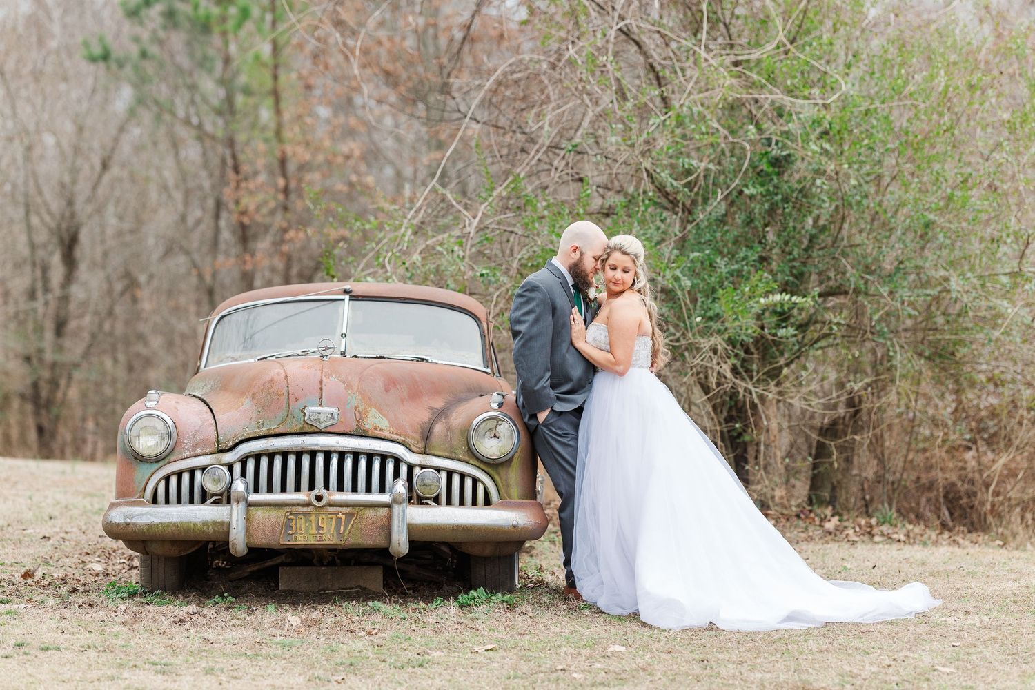 Romantic wedding photo with a rustic vintage car in a natural outdoor setting.