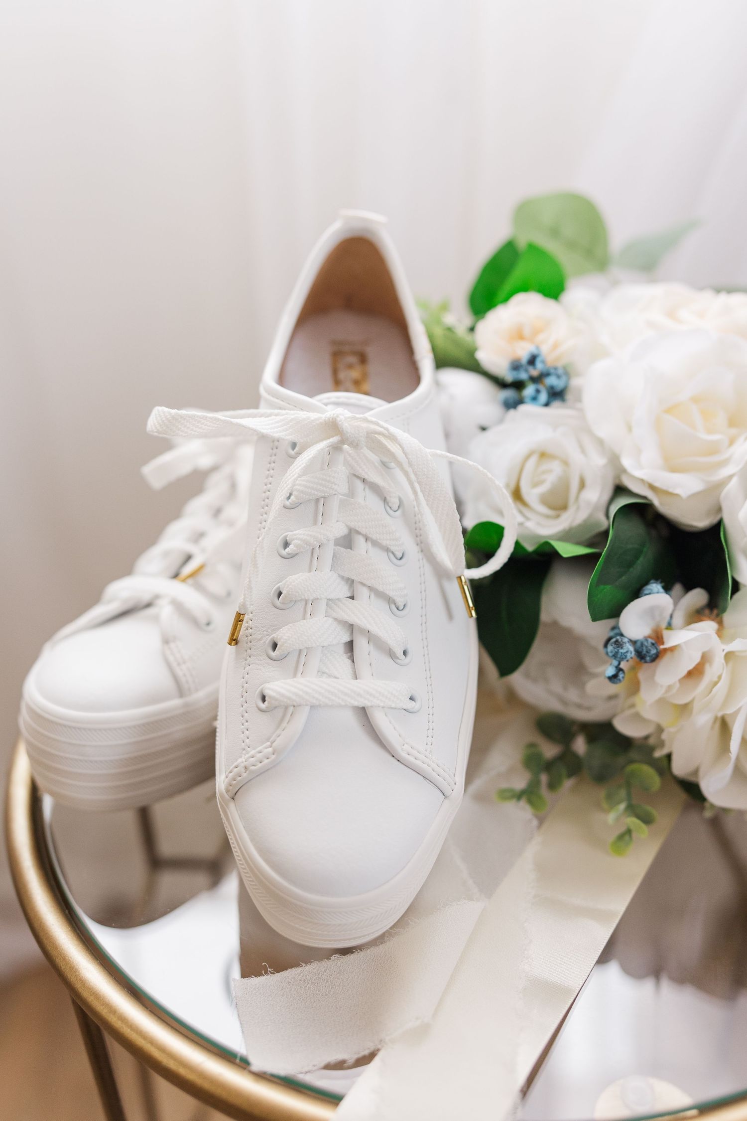 White canvas sneakers with ribbon laces displayed next to roses on a gold-rimmed table.