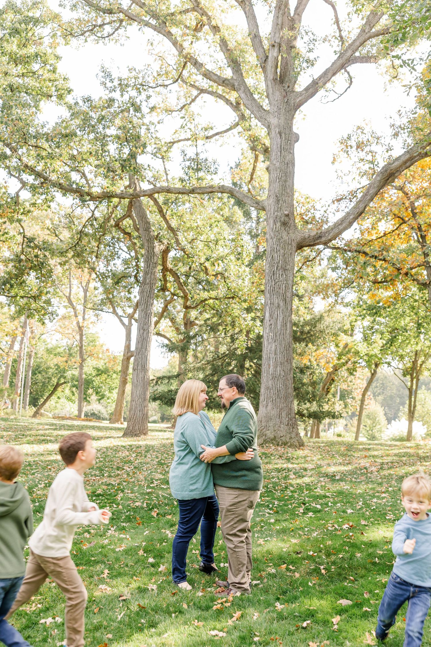 Family with children playing around parents embracing near large oak trees during autumn outdoor photo session.