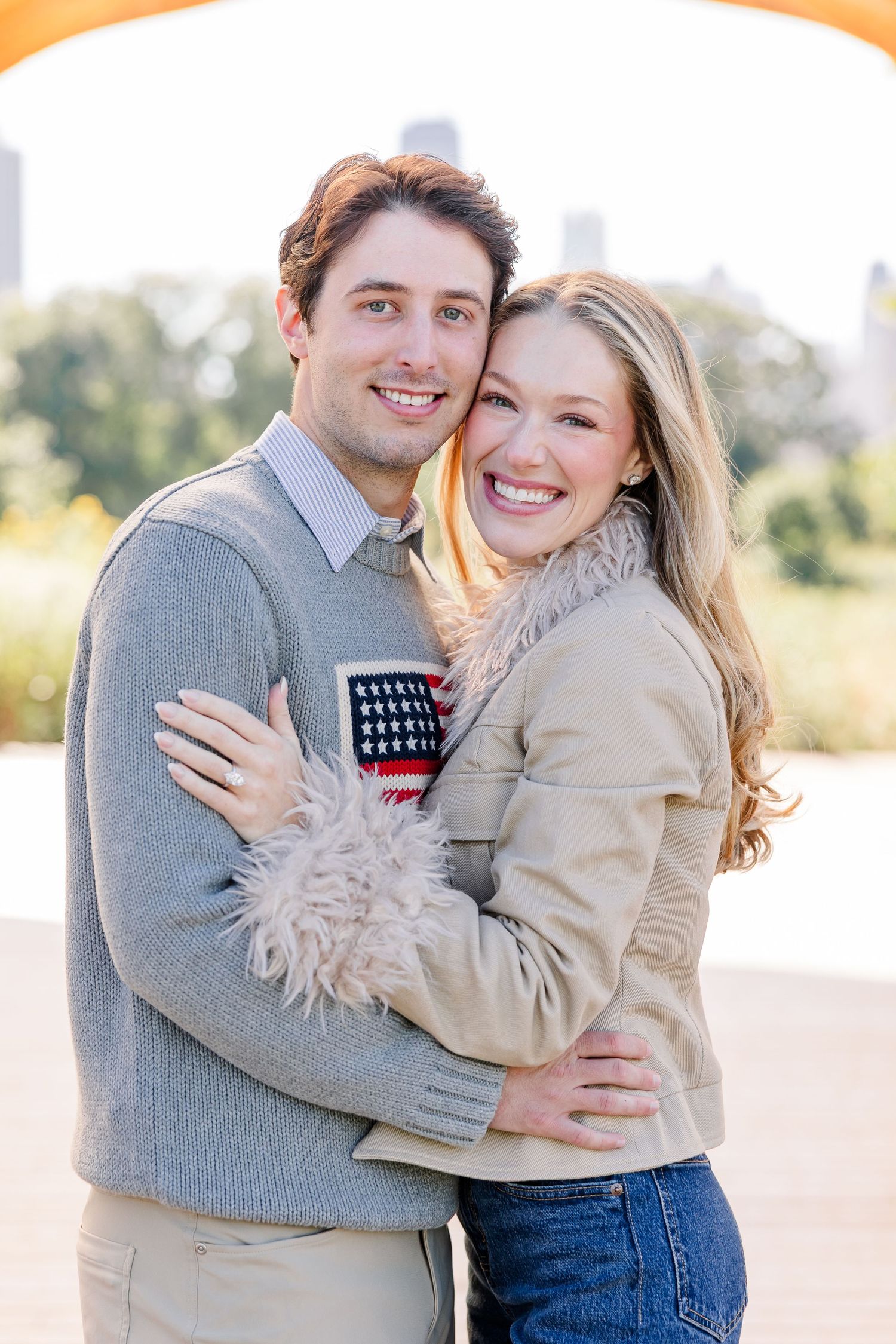 Smiling couple embracing outdoors, woman in feather-trimmed jacket and man in grey sweater with city views behind.