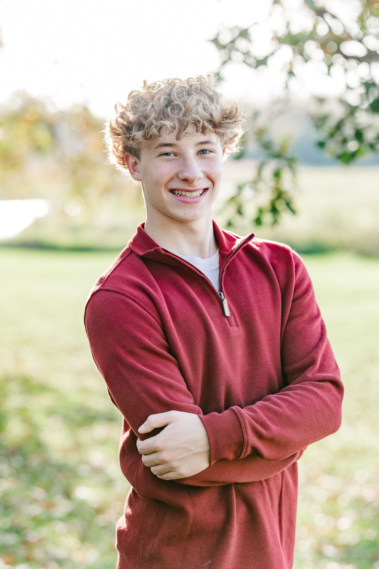 Smiling teenage boy with curly blond hair and a red quarter-zip pullover stands with arms crossed outdoors in warm autumn light.