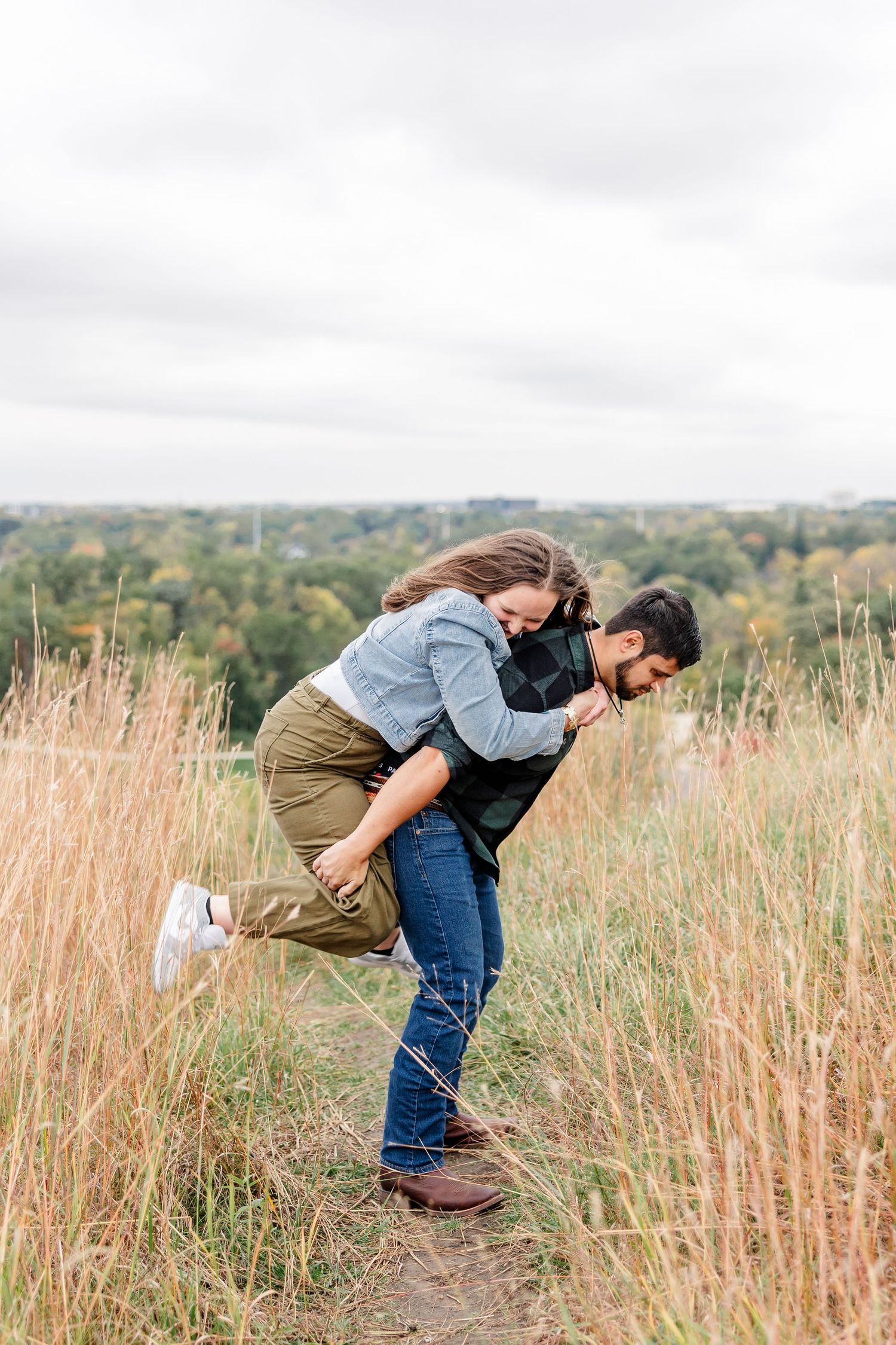 Man giving his partner a playful piggyback ride on a grassy hilltop trail with tall golden grass and a scenic view of fall trees.