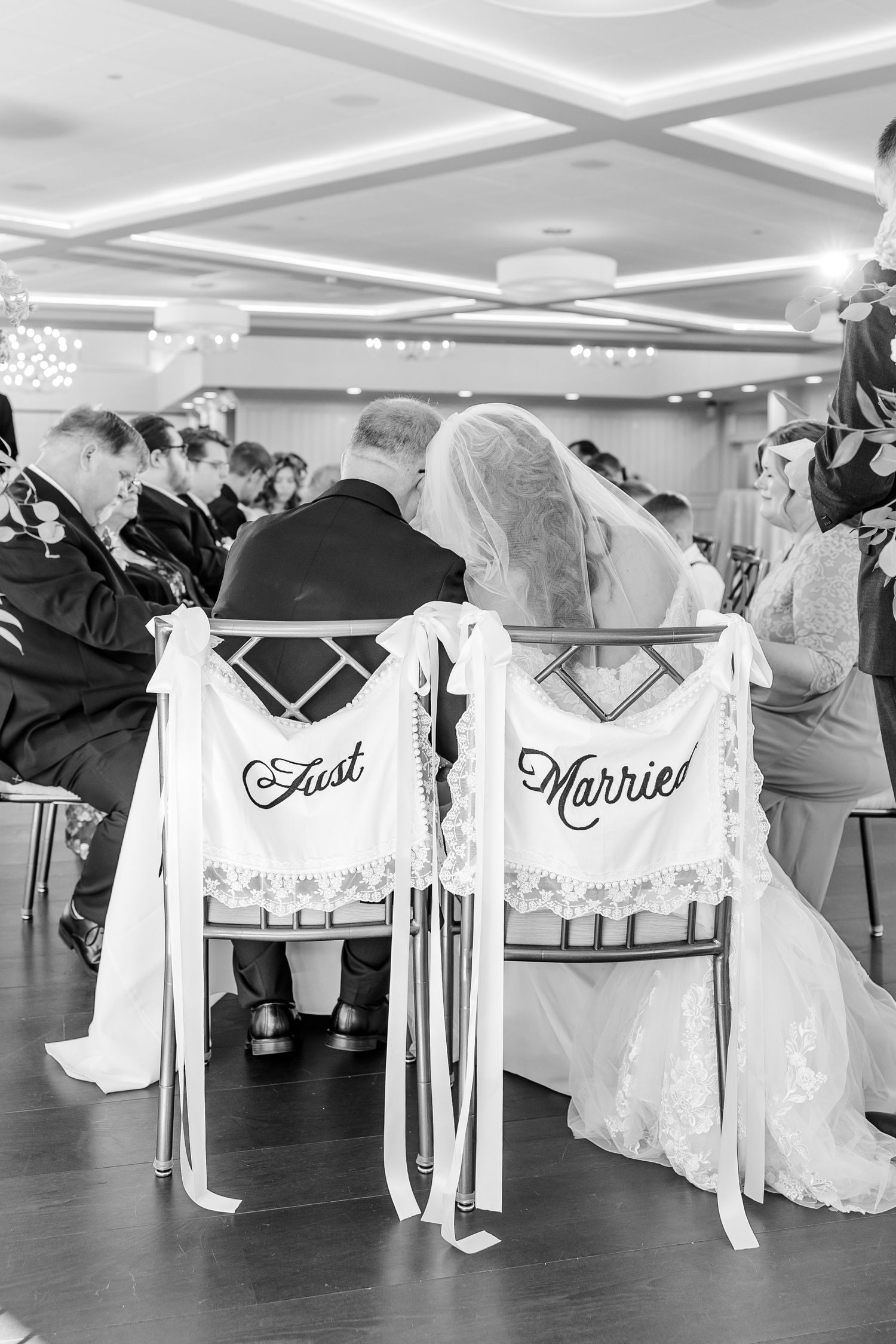 Black and white photo of a bride and groom seated from behind at their ceremony, chair signs reading Just Married.