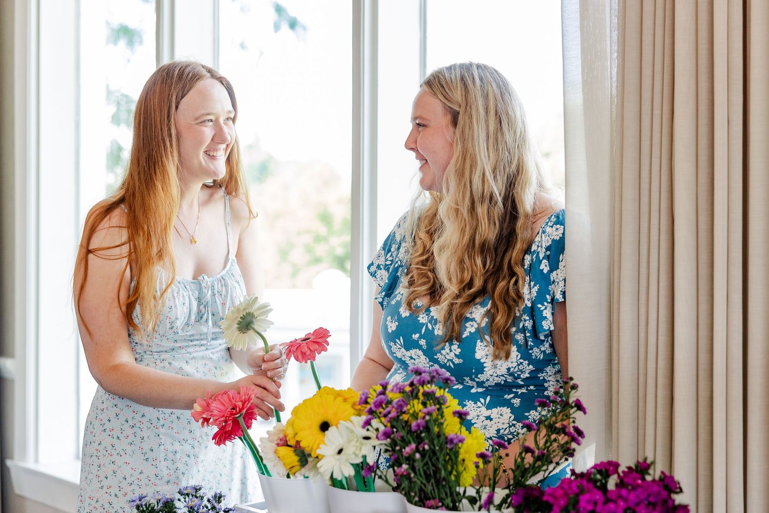 Two women laughing together while arranging colorful flowers near a bright window.
