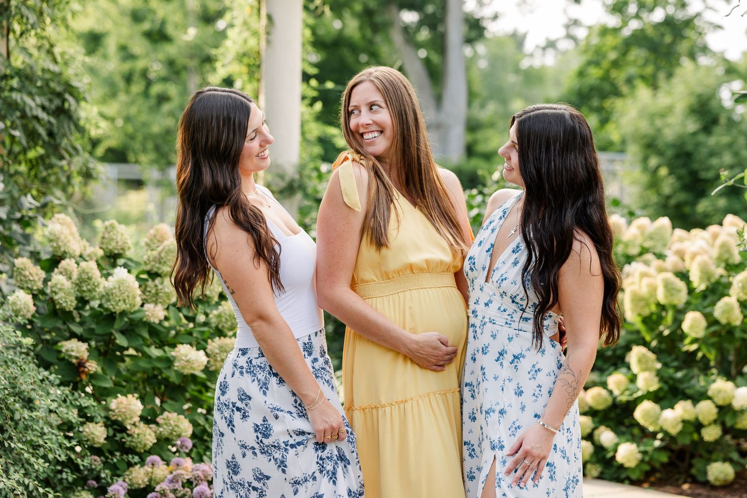 Three women smiling together in a garden, center woman in yellow dress holding baby bump.