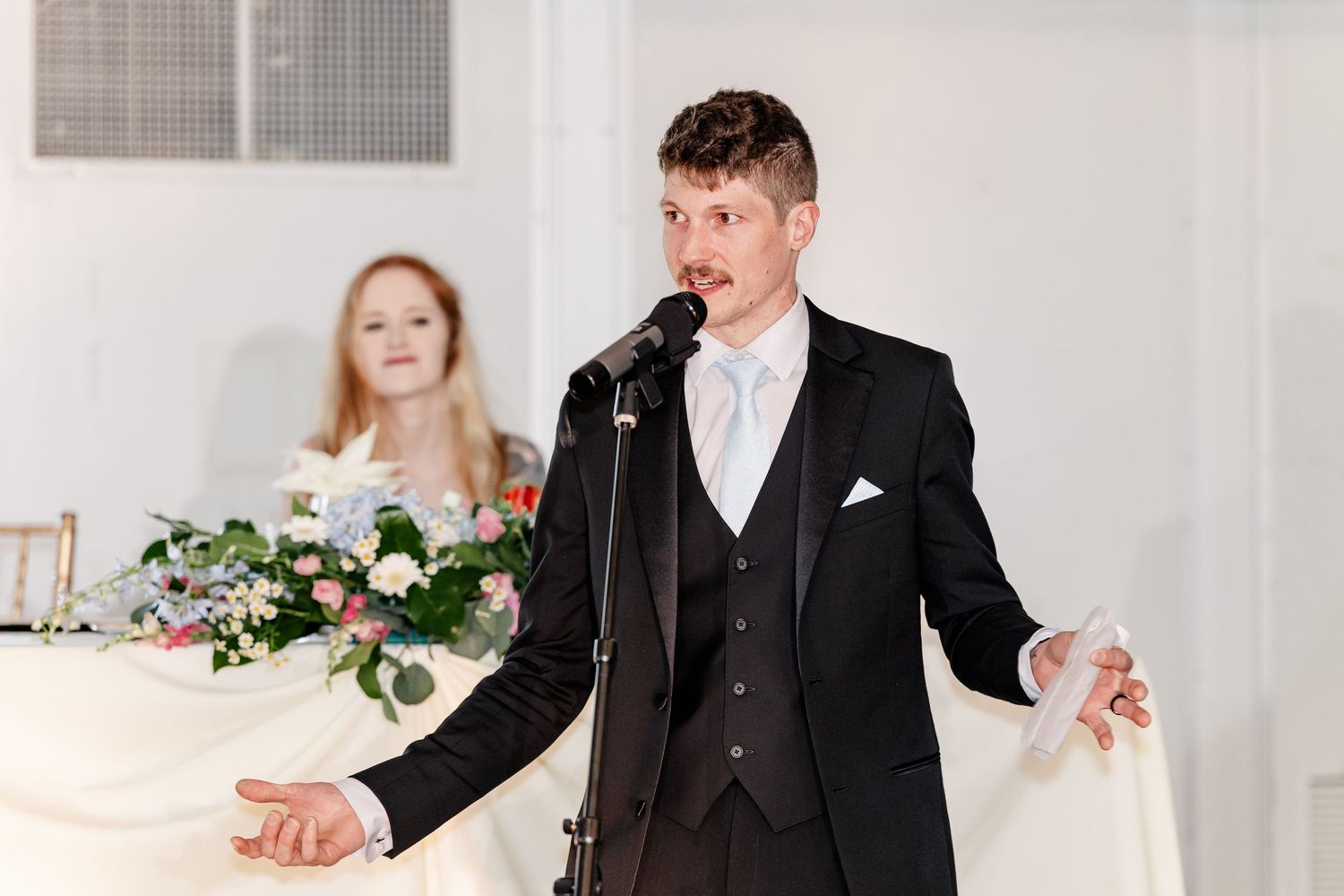 Man in black tuxedo giving speech at microphone during wedding reception with bride in background.