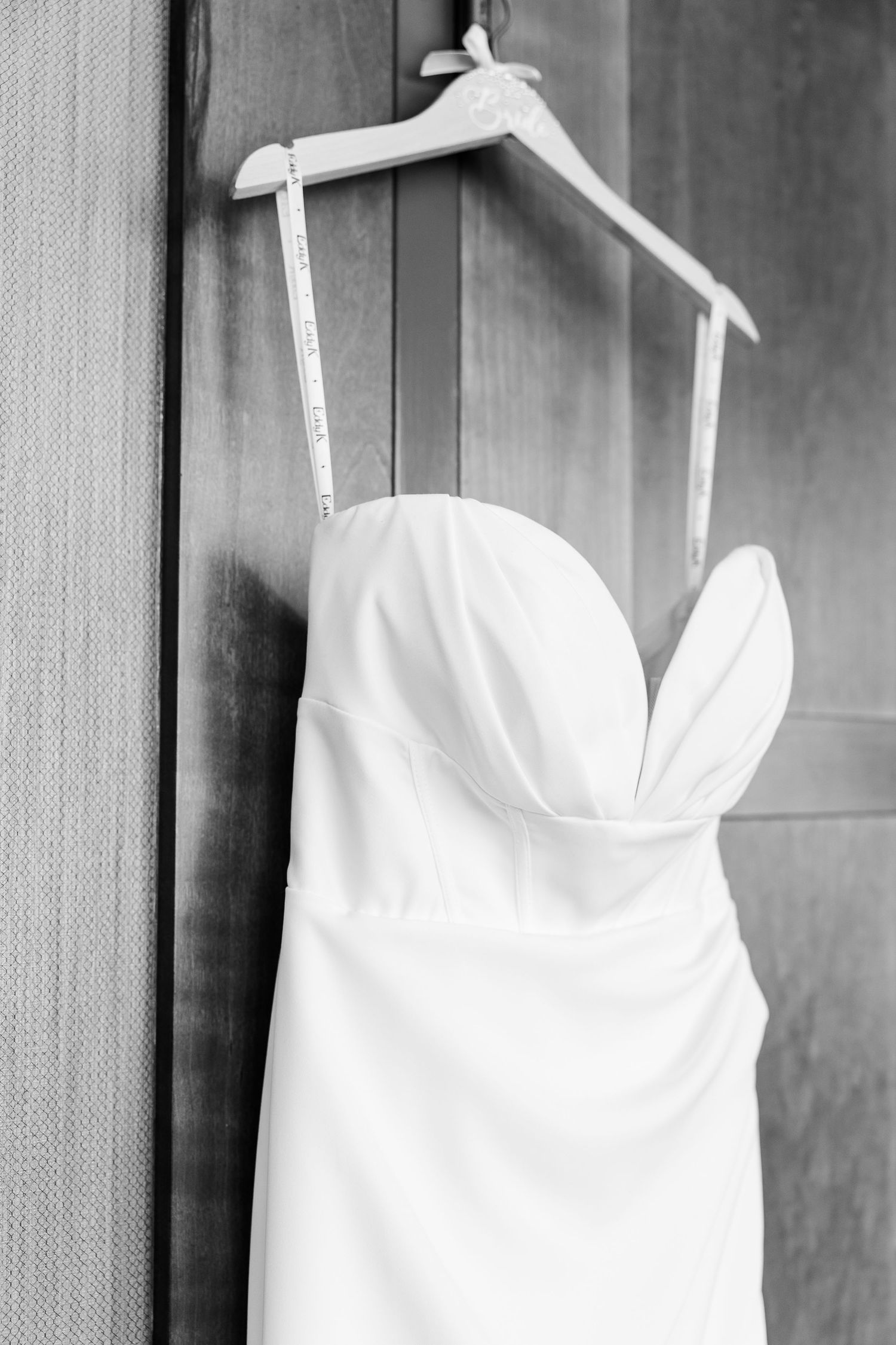 A strapless white wedding gown hanging on a wooden hanger against a dark door in black and white.