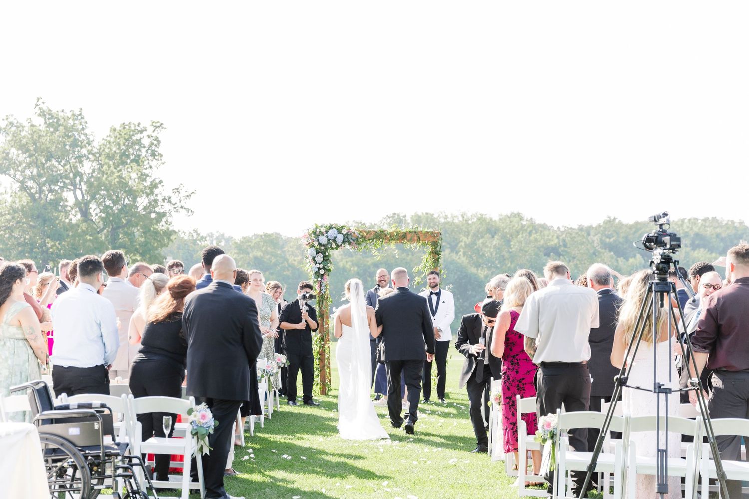 Outdoor wedding ceremony with floral arch by waterfront as couple exchanges vows.