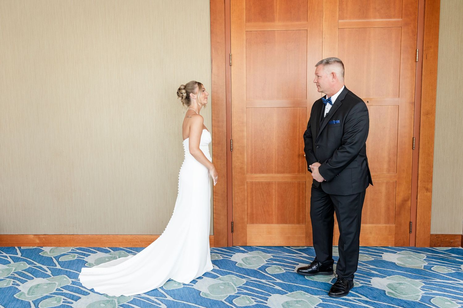 Bride in white gown shares first look moment with groom in black suit in hotel hallway.