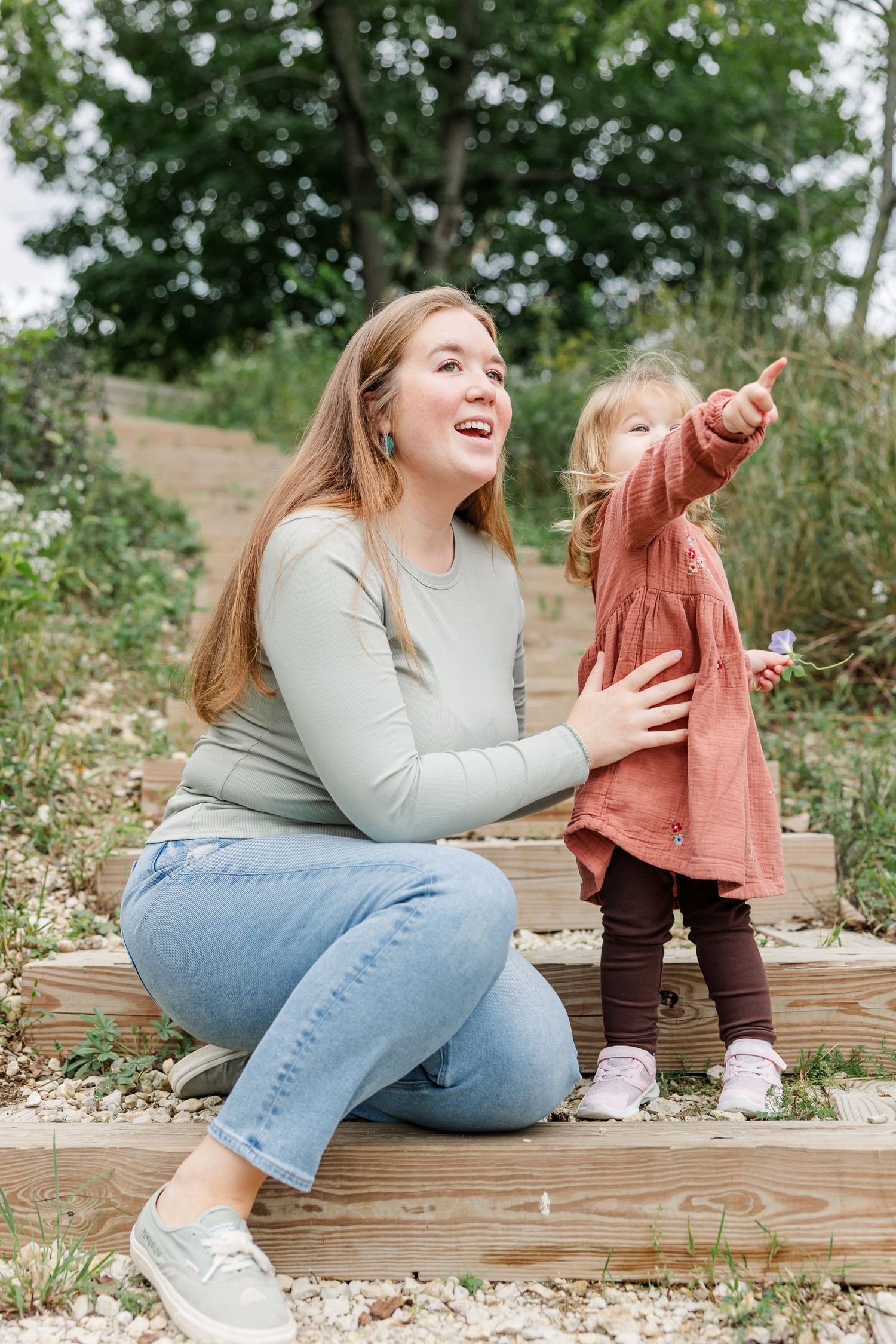 A laughing woman sits on wooden steps outdoors while a toddler in a pink dress points excitedly at something nearby.