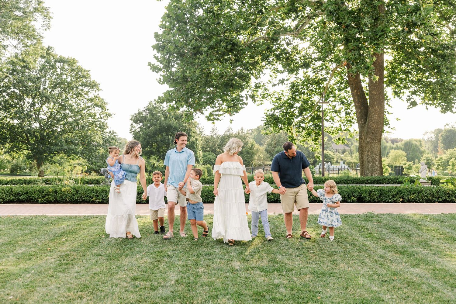 A large family group poses together on a lush green lawn surrounded by manicured hedges and trees on a sunny day.