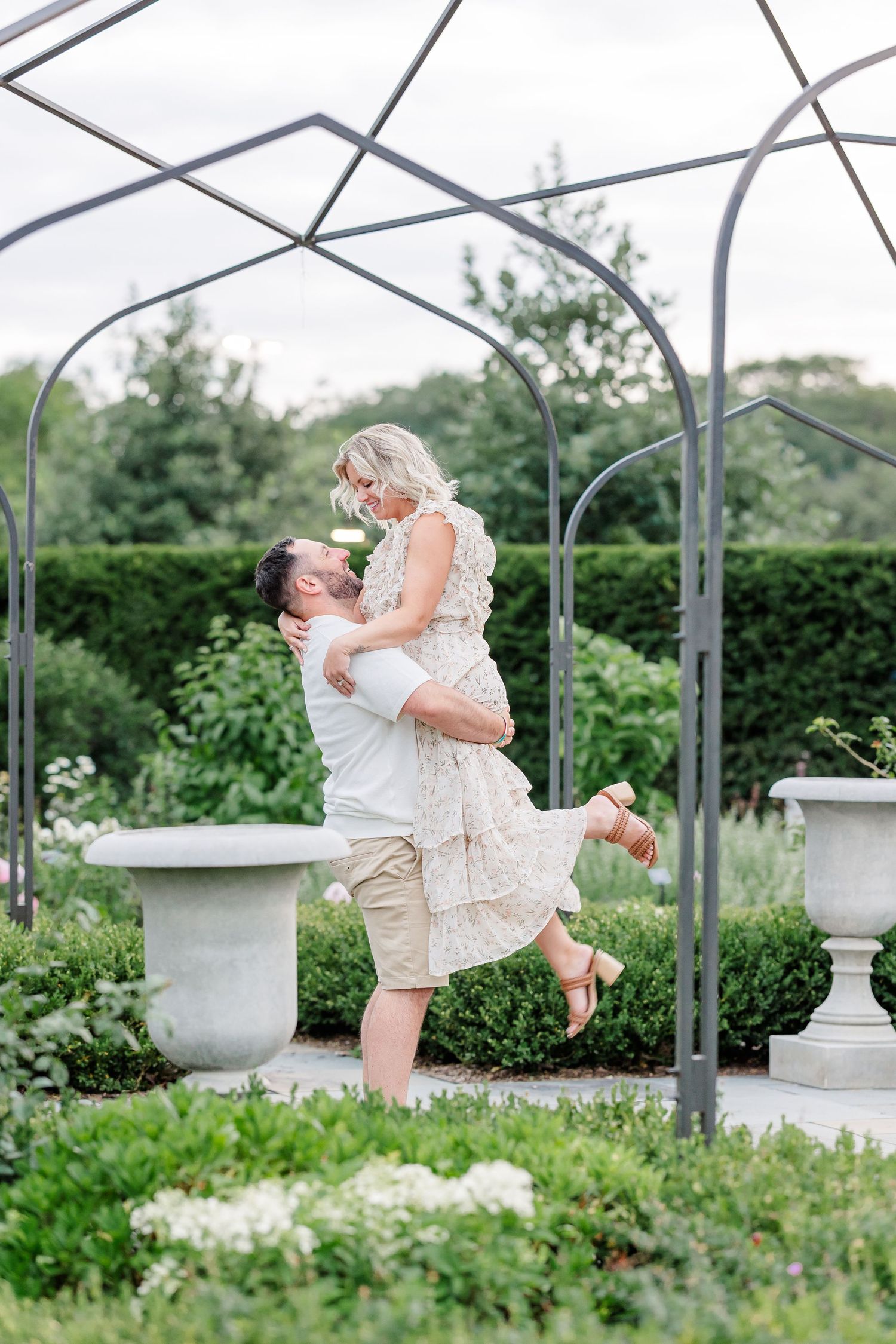 A man lifts a smiling blonde woman under a metal garden arch surrounded by lush green hedges.
