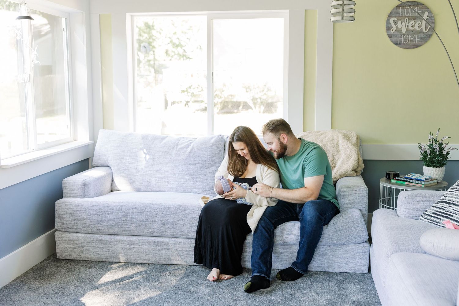 A couple sits on a gray couch admiring their newborn baby in a bright, cozy living room.