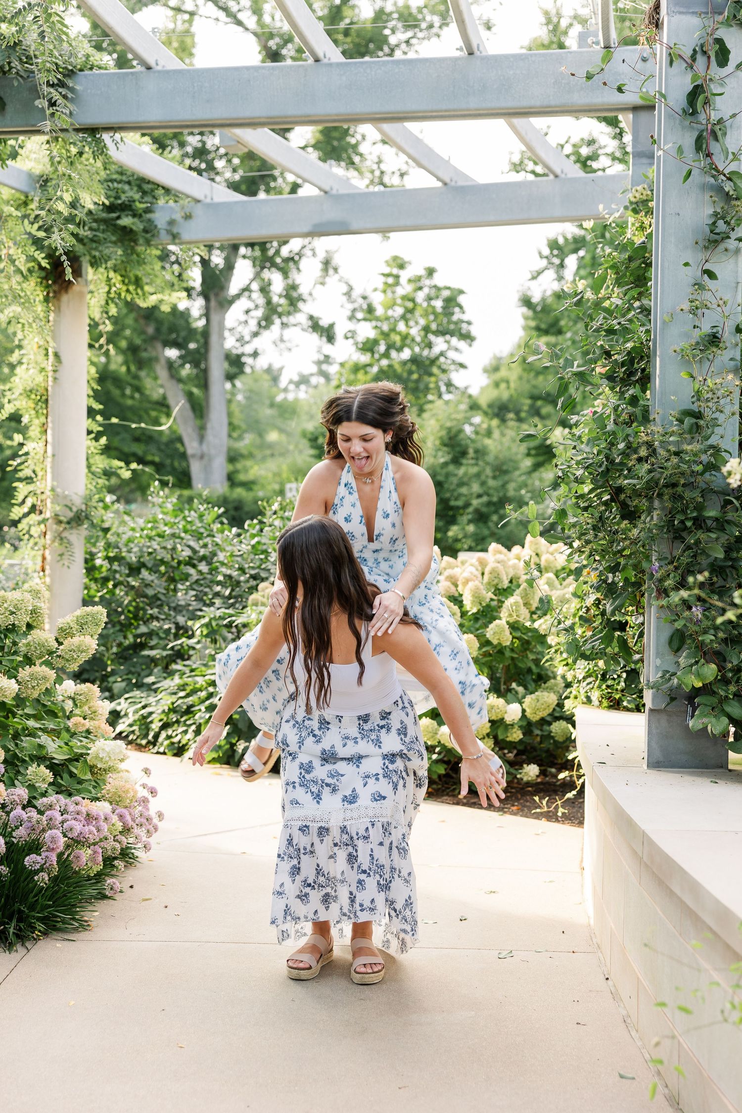 Woman in white floral dress laughs joyfully with a young girl under a pergola surrounded by blooming hydrangeas.