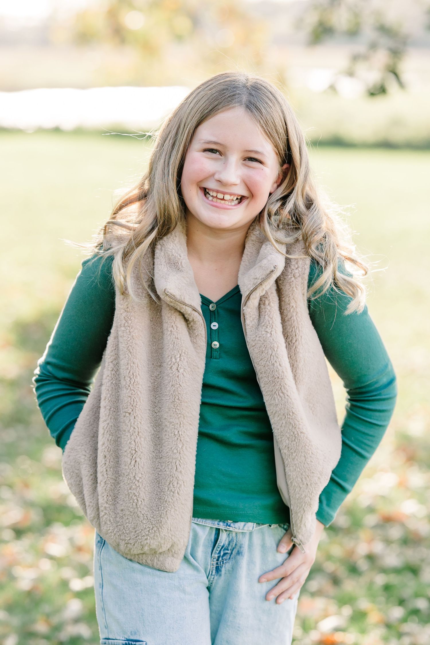 A smiling girl with long wavy hair wears a green top, tan fuzzy vest and light jeans while posing outdoors on a leaf-covered lawn.