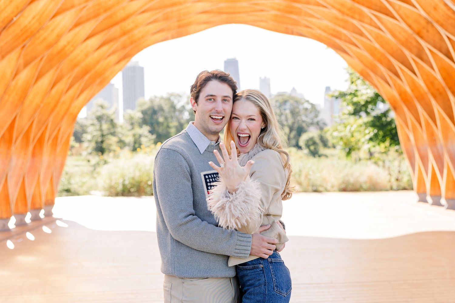Happy couple embracing under orange arch structure in Chicago park, woman showing engagement ring, city skyline in background.