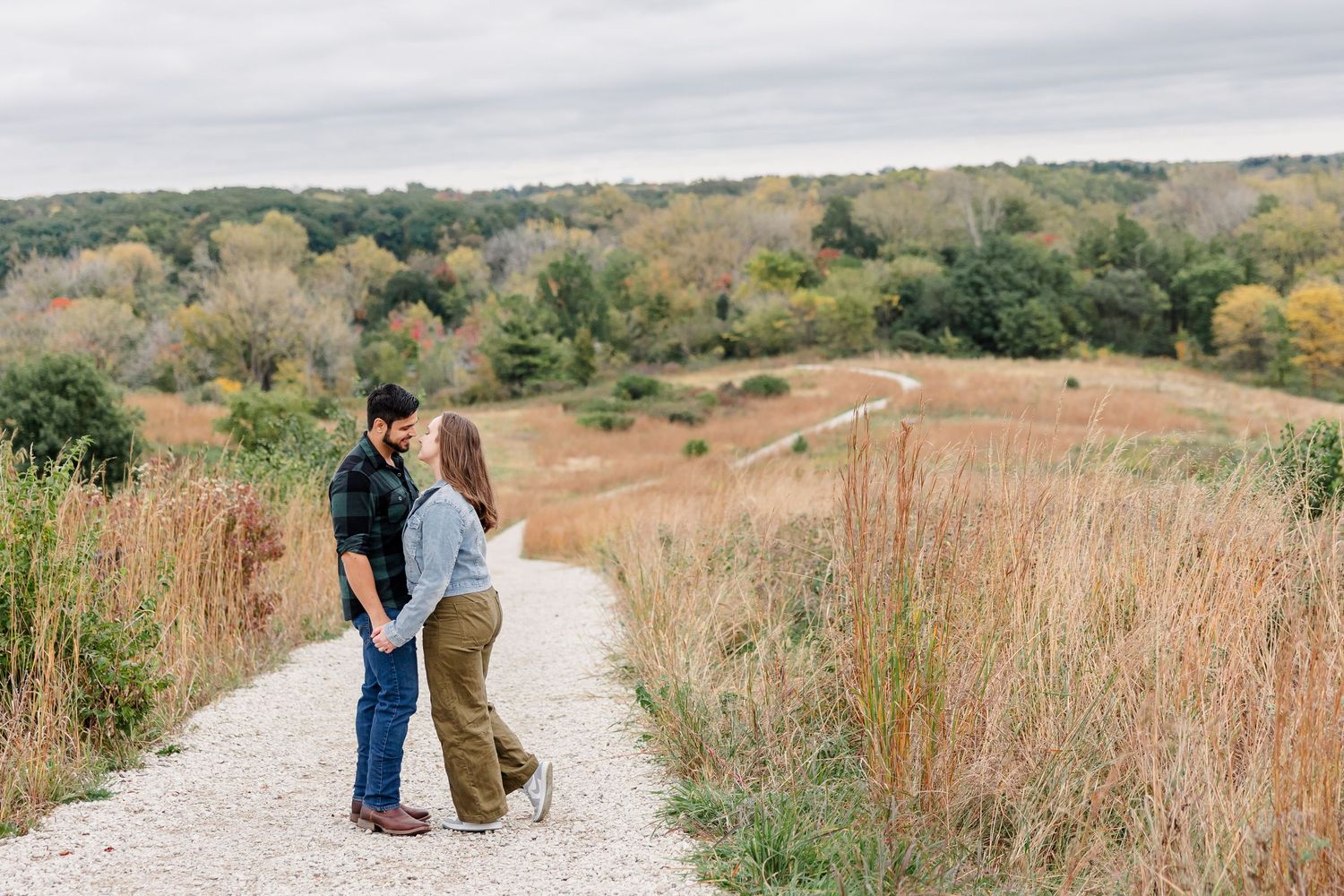 Couple embraces on a gravel path winding through tall prairie grass and rolling hills with colorful autumn trees in the distance.