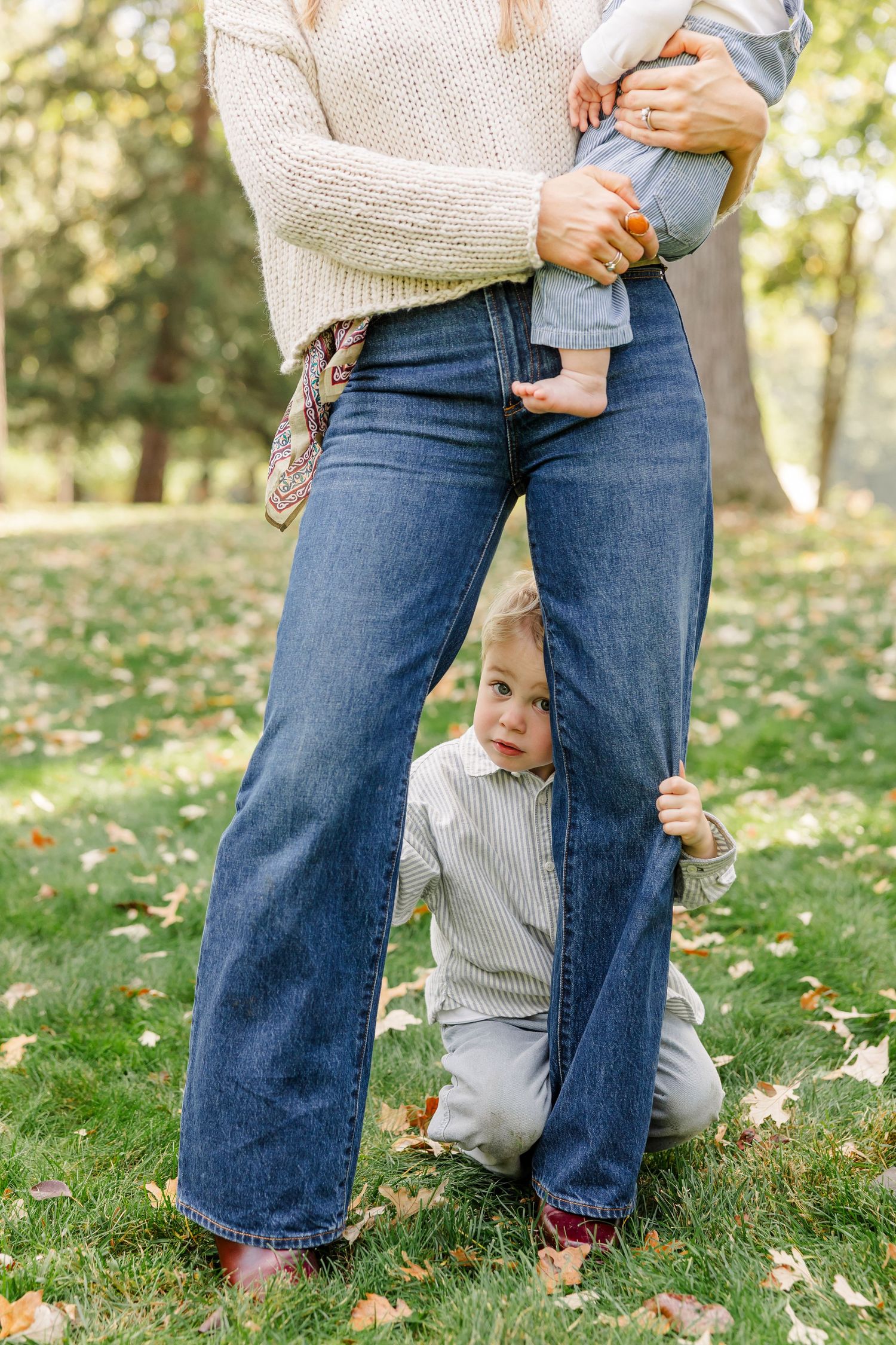 Young toddler peeks through mothers legs while she holds a baby outdoors.