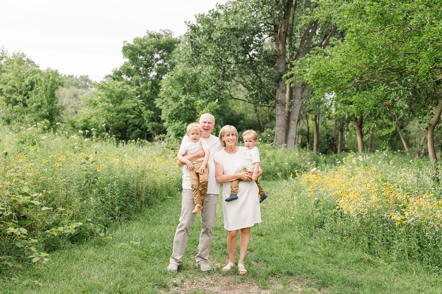 Grandparents holding two young grandchildren on a scenic wildflower meadow path.