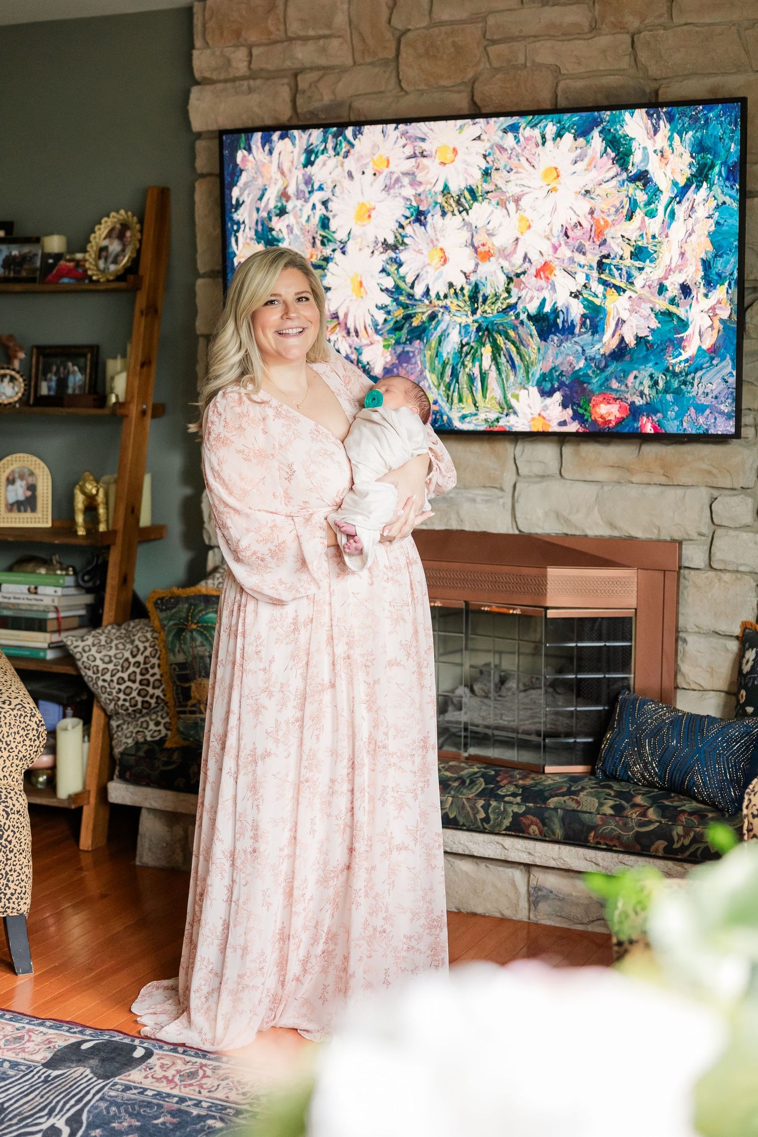 Smiling blonde mother in pink floral gown holding newborn baby in front of stone fireplace with floral artwork.