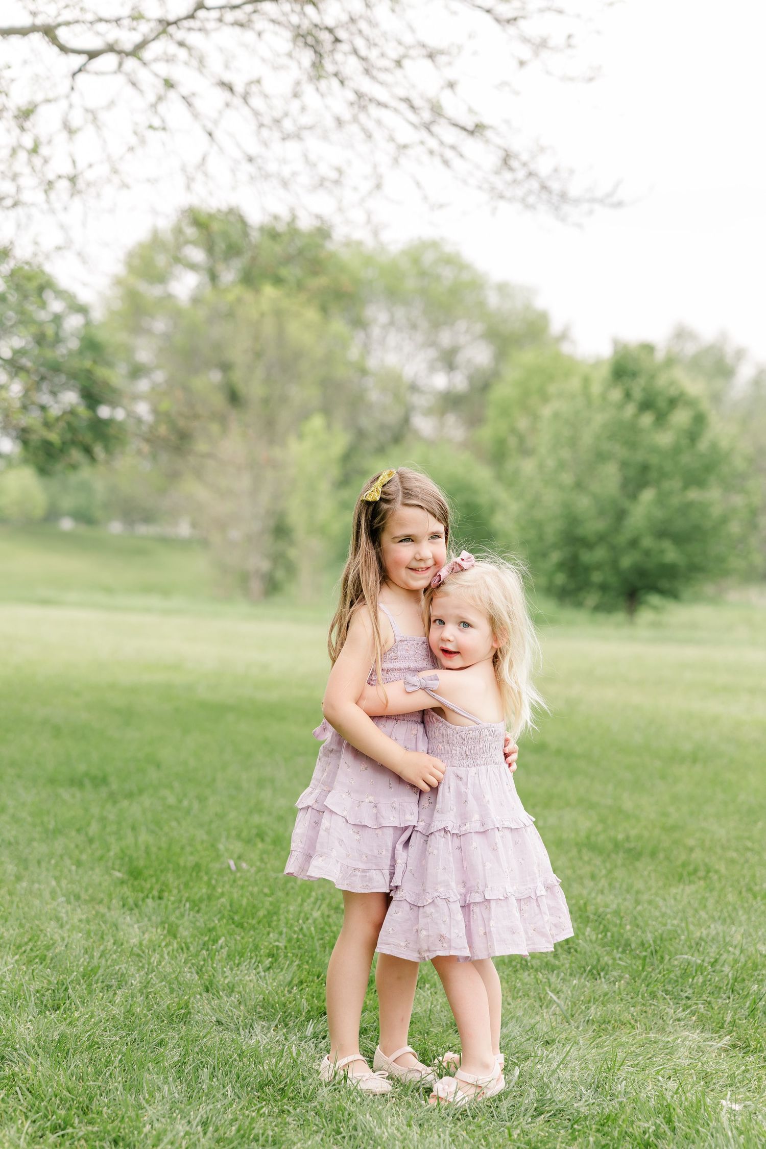 Two young sisters in matching lavender ruffle dresses hug each other while standing in a green field.