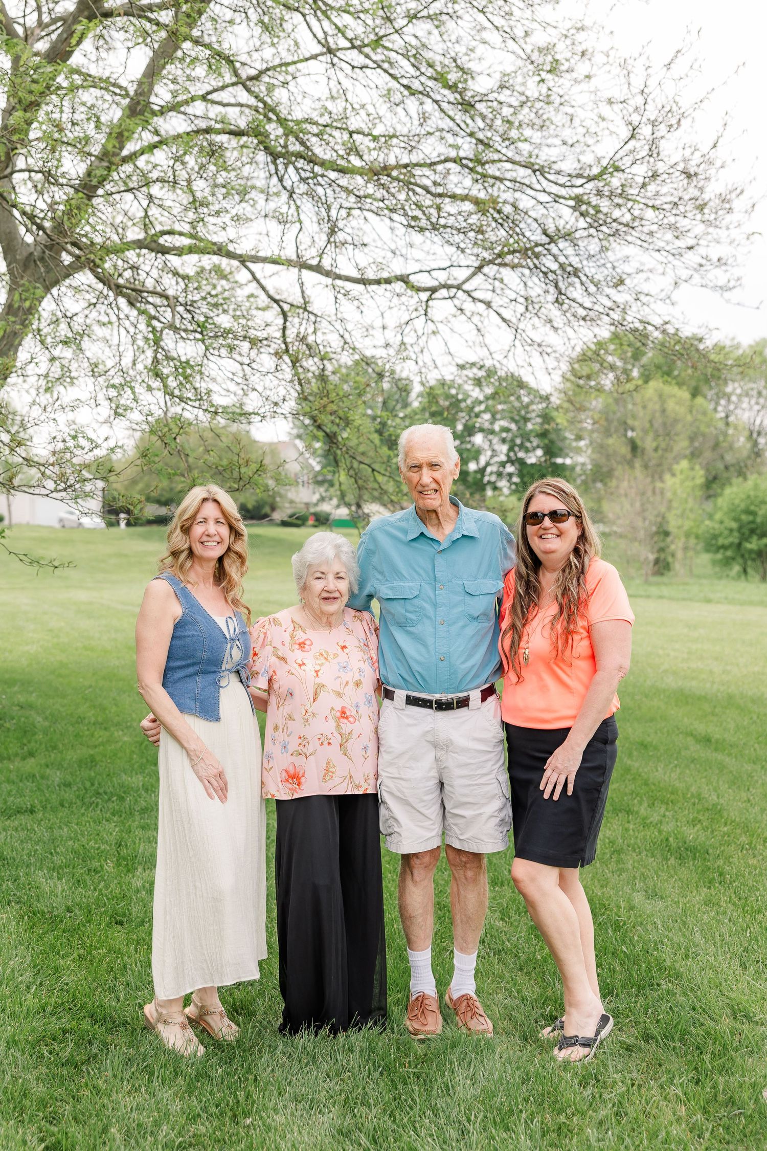 Four generations of family posing together outdoors on green grass beneath a large tree on a sunny spring day.