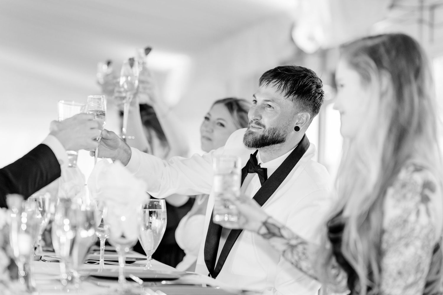 Black and white photo of wedding guests raising champagne glasses in a toast at a reception dinner table.