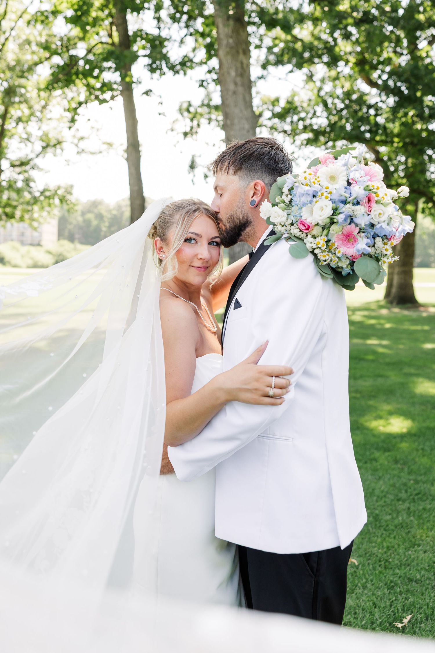 Bride and groom embracing under trees, veil flowing, holding colorful wedding bouquet.