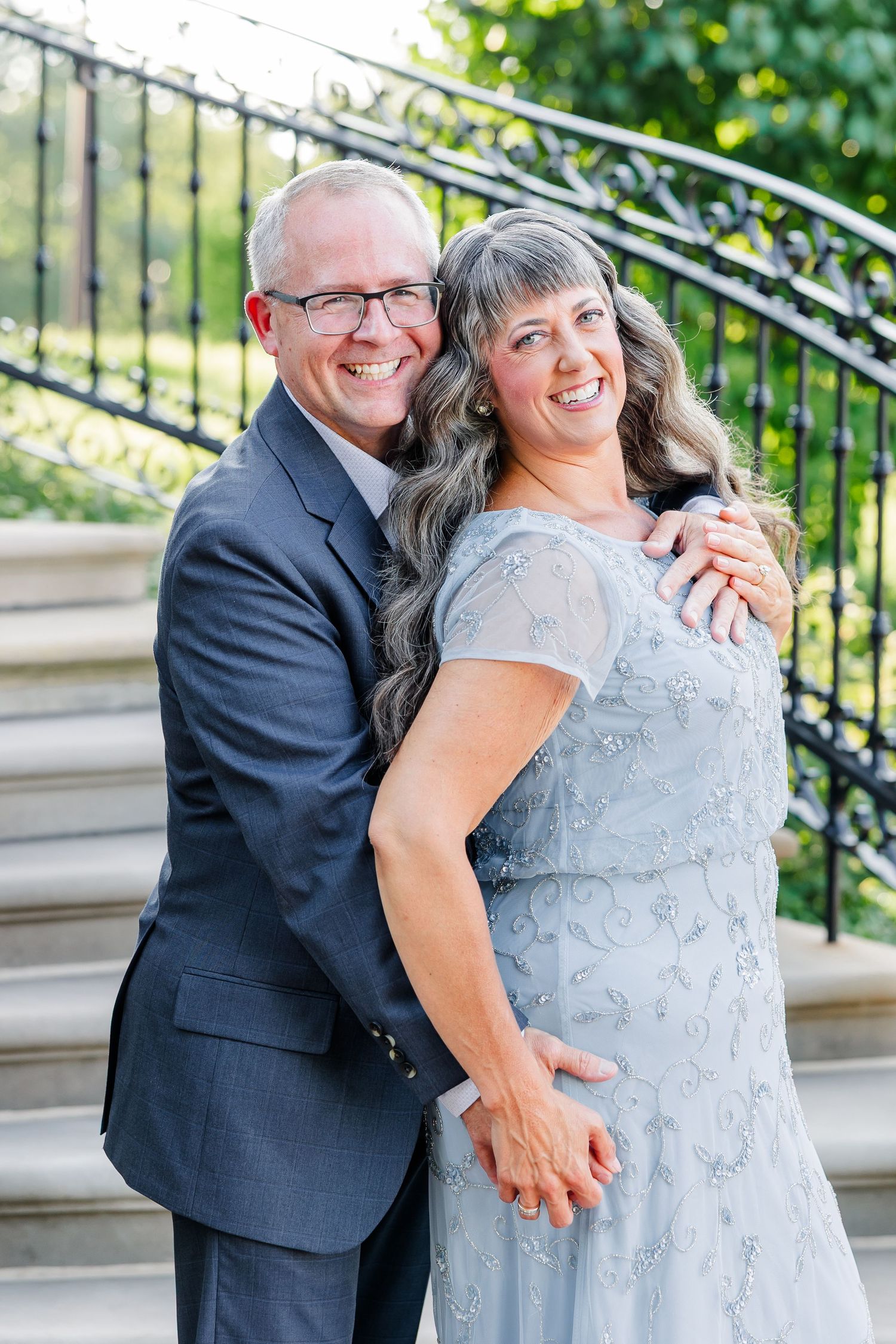 Smiling couple in formal attire poses on outdoor steps, man in gray suit embraces woman in light blue beaded gown.